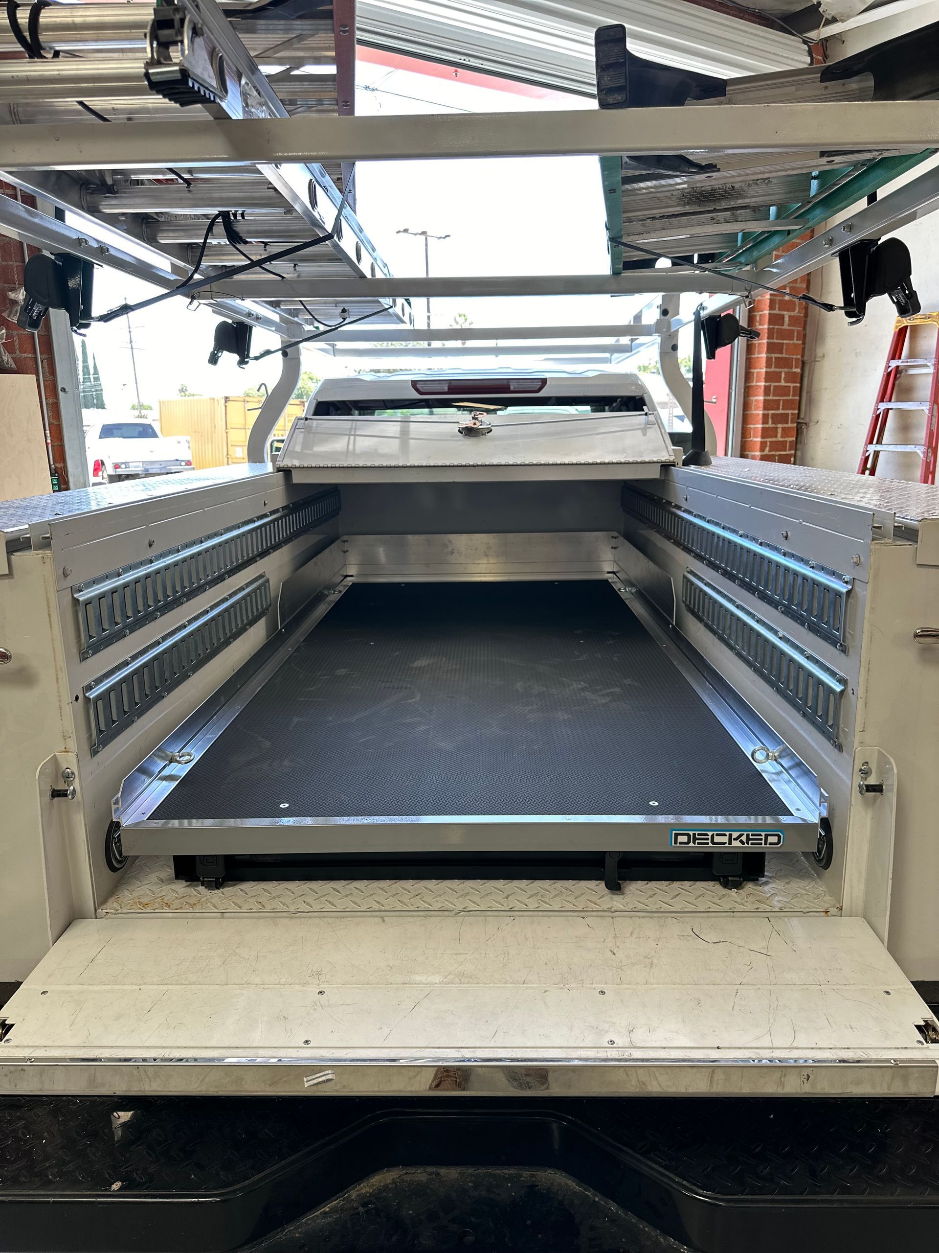Open cargo bed of a service truck featuring a central pull-out metal sliding tray and overhead ladder racks.