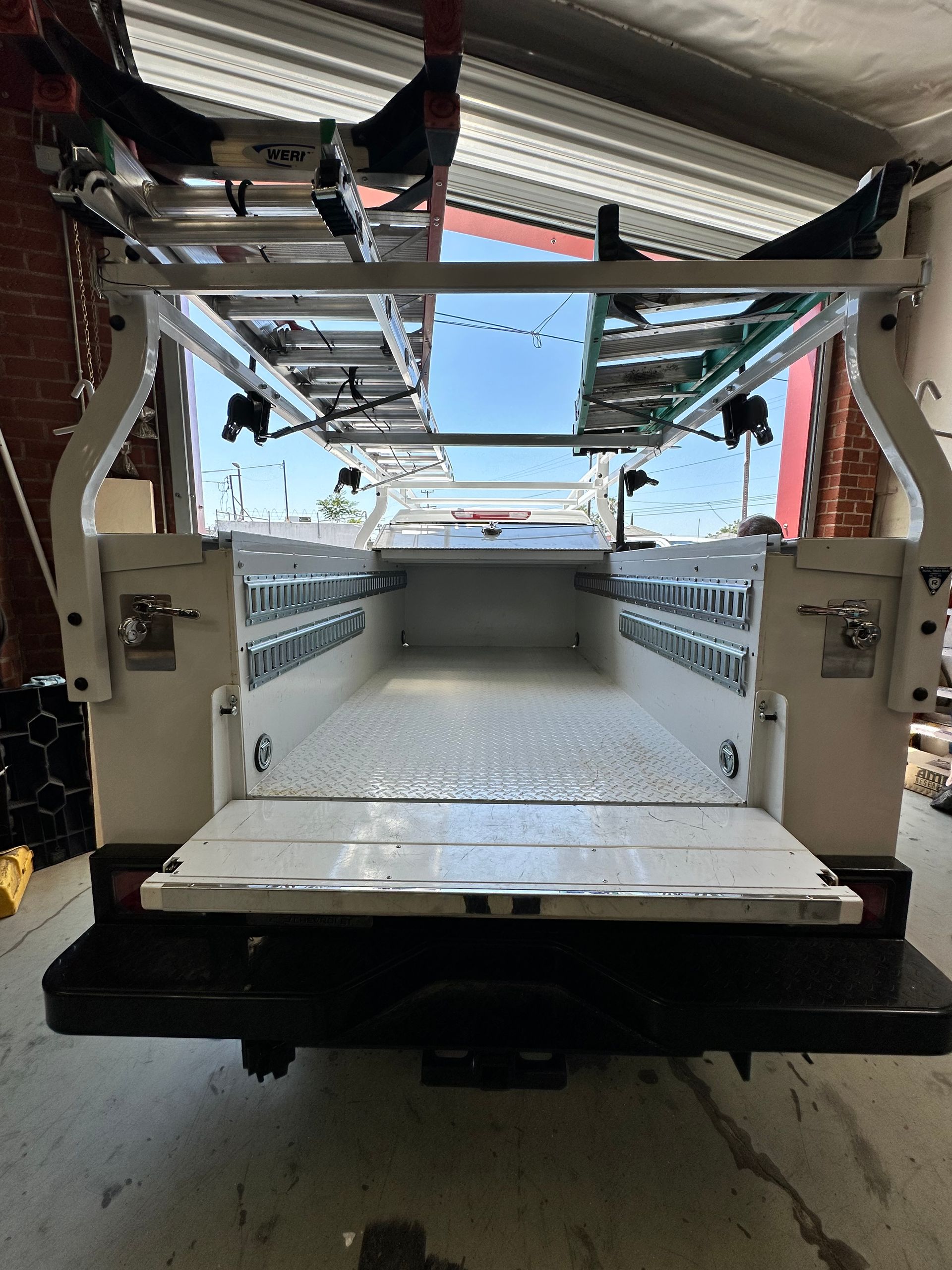 The open bed of a white utility service truck with an overhead ladder rack, viewed from the rear inside a garage.