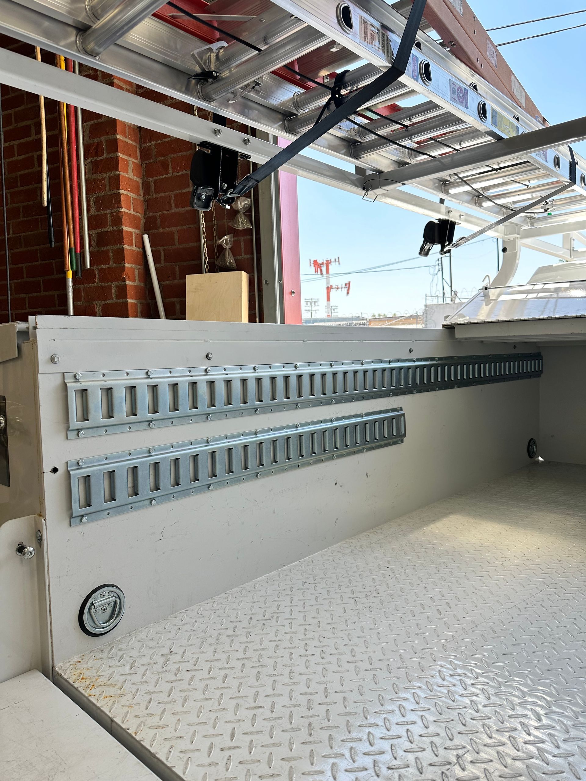 Inside a service truck bed with metal tracks on the wall, a diamond-plate floor, and a ladder rack mounted above.