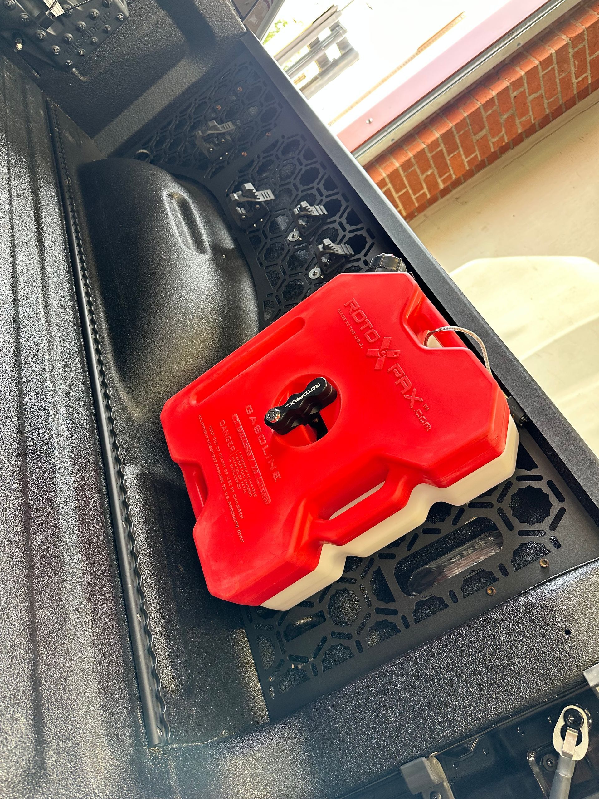 A red Rotopax fuel canister is securely mounted onto the textured black metal panel of a truck bed.