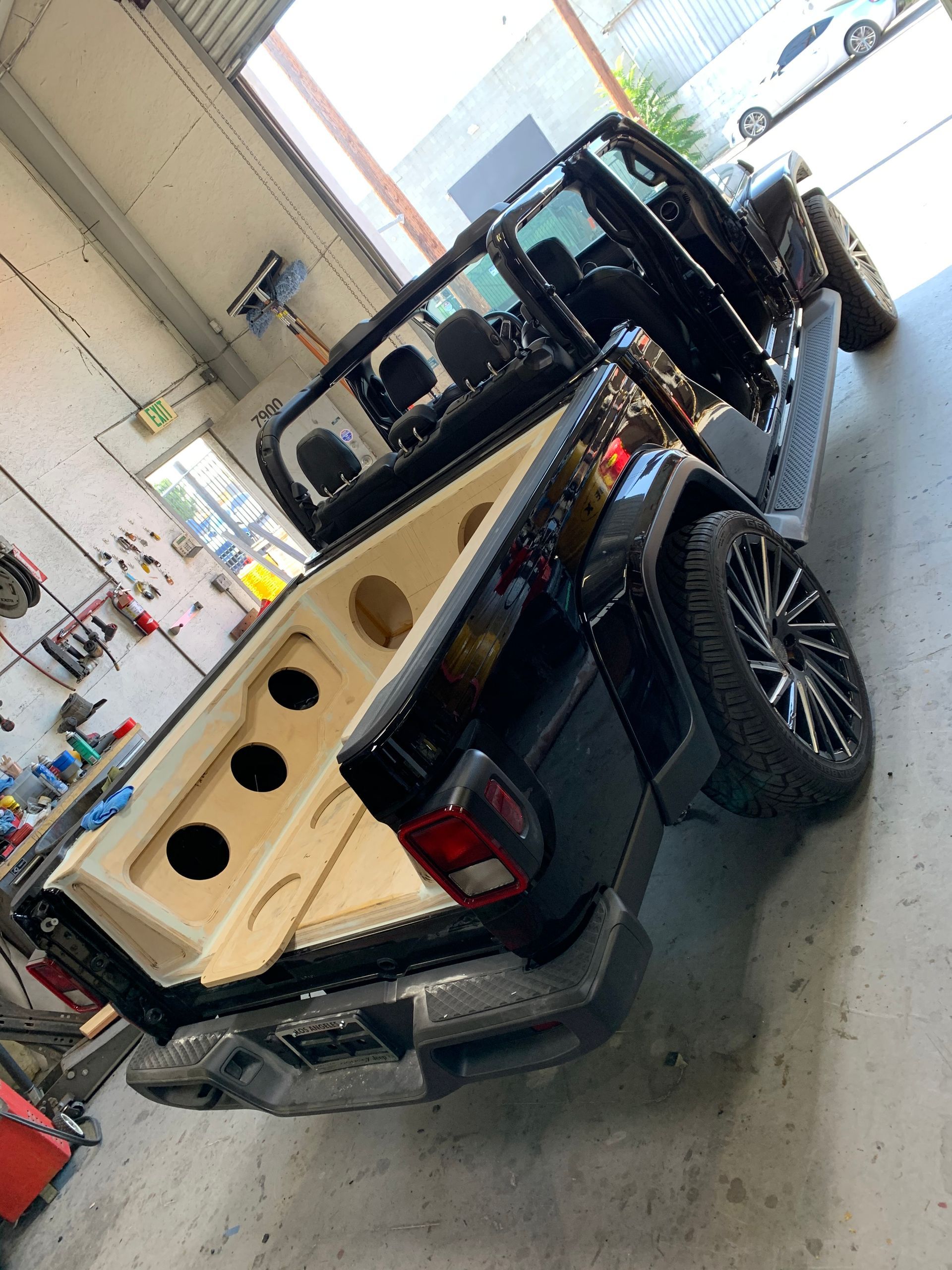 A black Jeep Gladiator with a custom wooden speaker enclosure installed in the cargo bed inside a workshop.