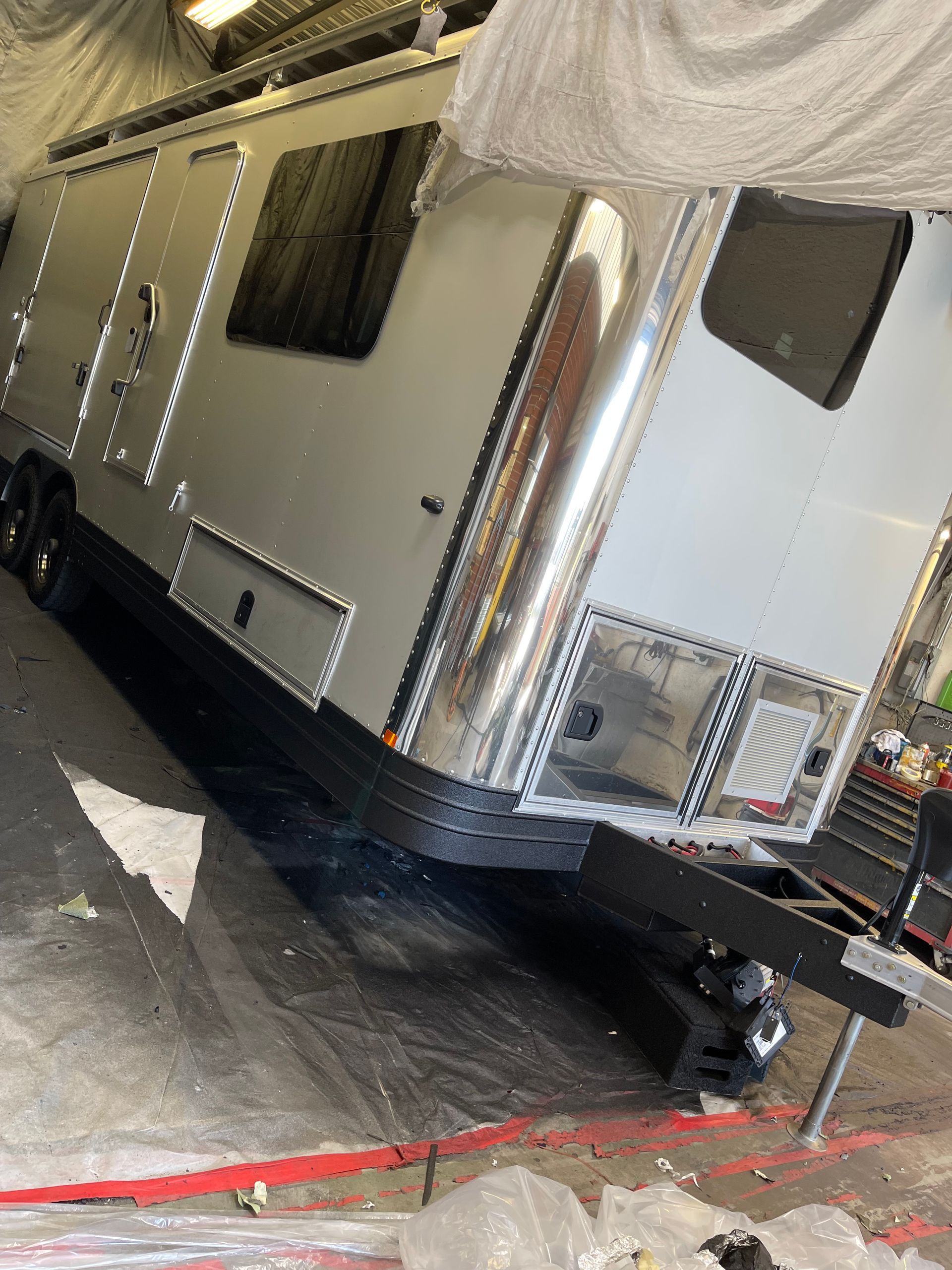 A silver-paneled metal trailer with a black frame, parked inside a workshop with white draped covers in the background.