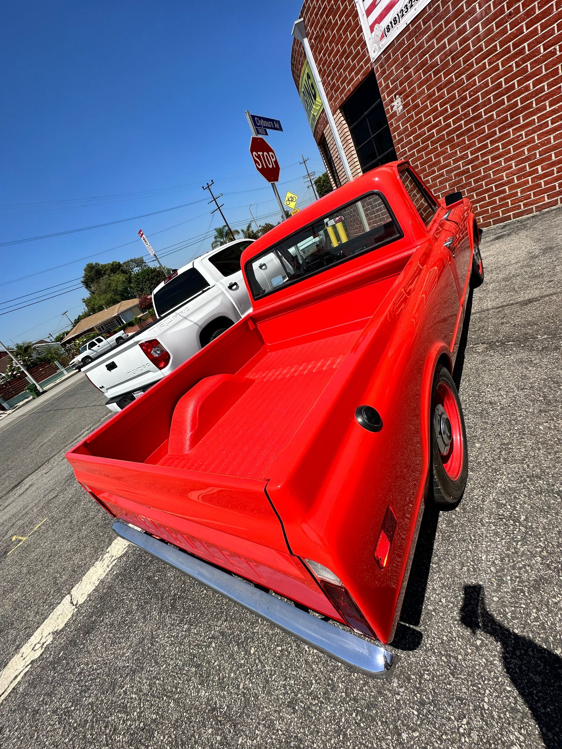 A bright red vintage pickup truck parked on an asphalt lot beside a brick building on a sunny day.