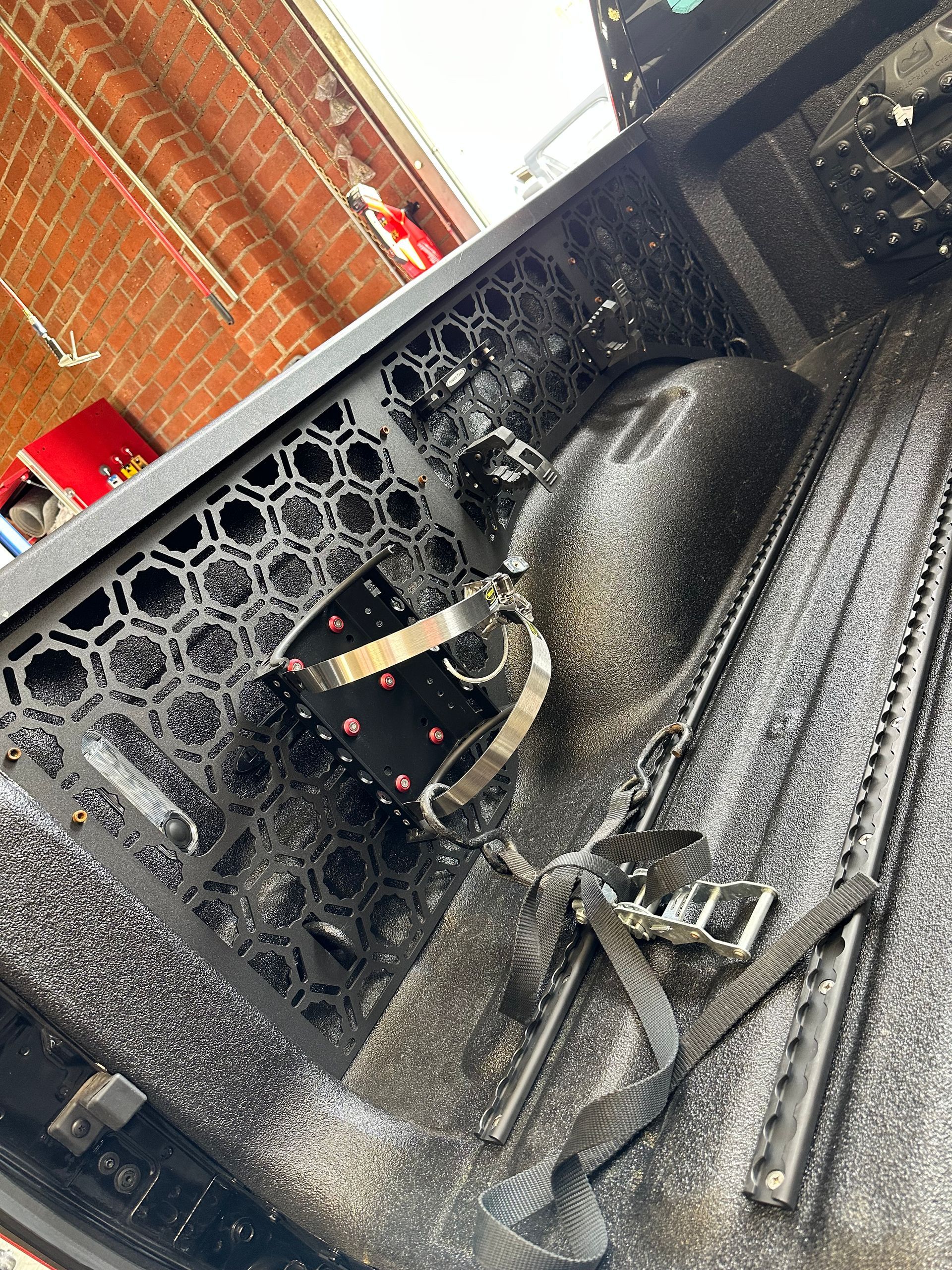 A black truck bed interior with a honeycomb-patterned side panel, a black metal mount, and a black ratchet strap.