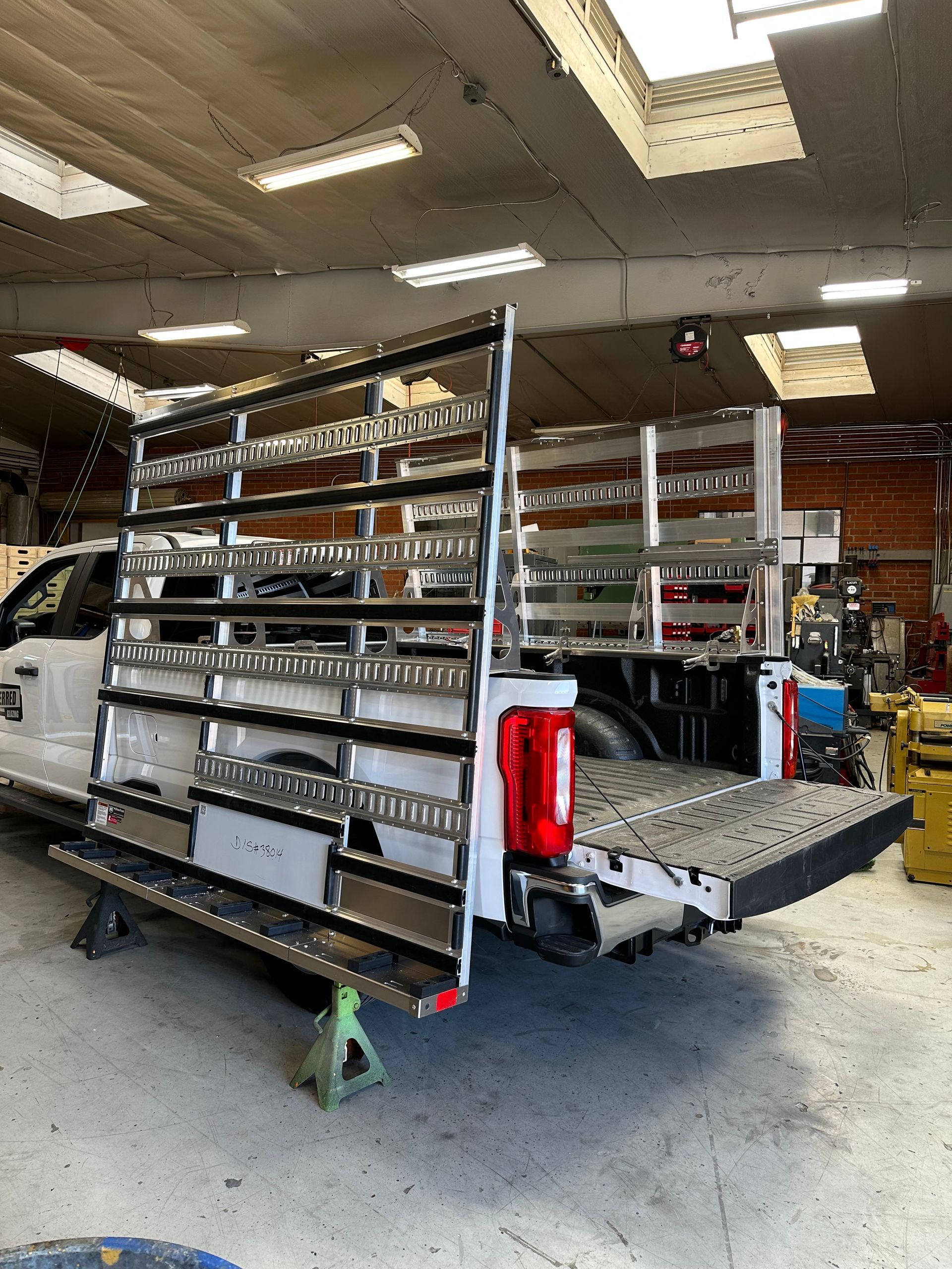 A white truck bed equipped with a custom aluminum glass rack sits on jack stands inside a workshop.