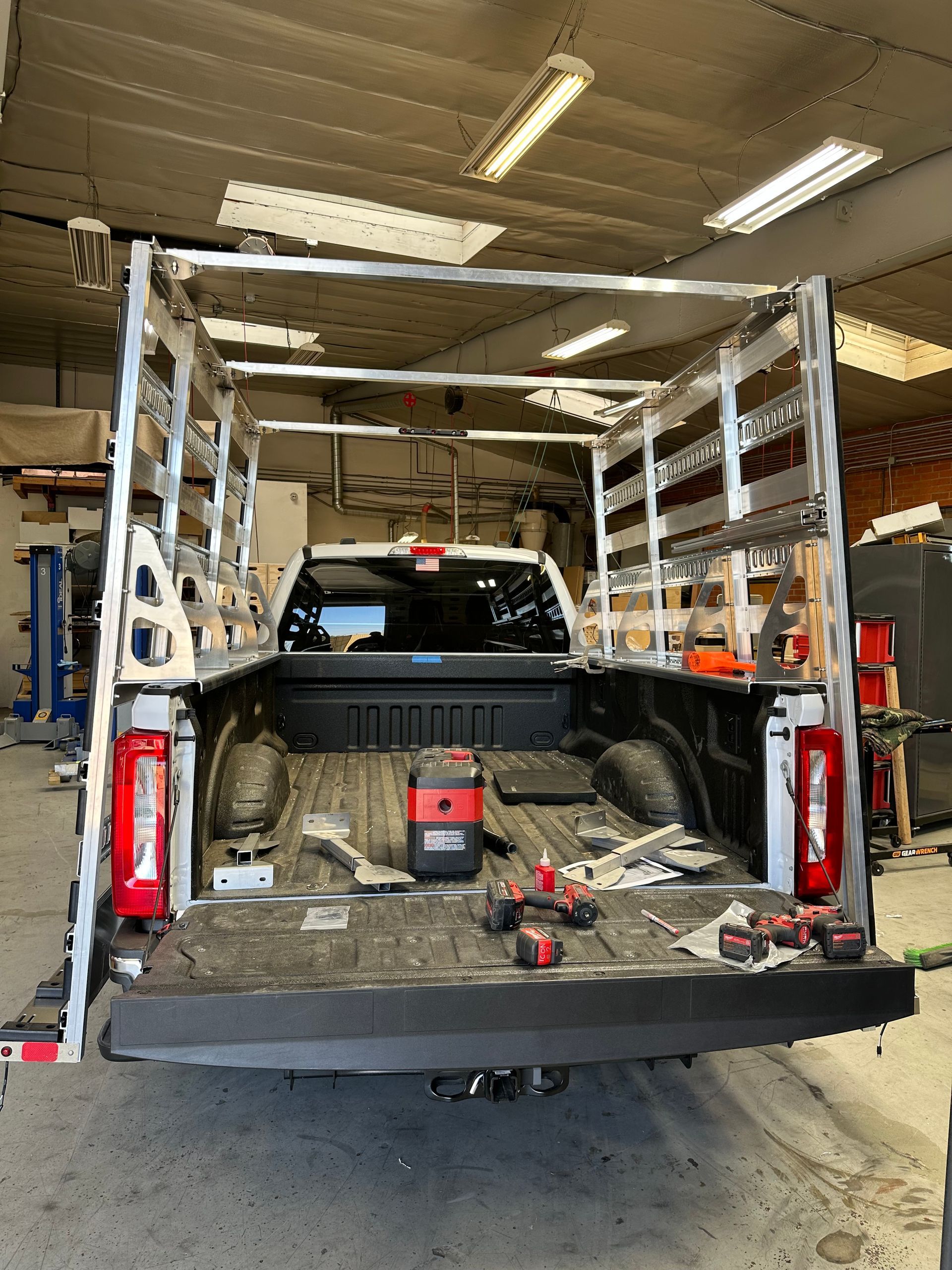 A white pickup truck in a garage with a tall, metal cargo rack installed over the bed, holding tools on the tailgate.