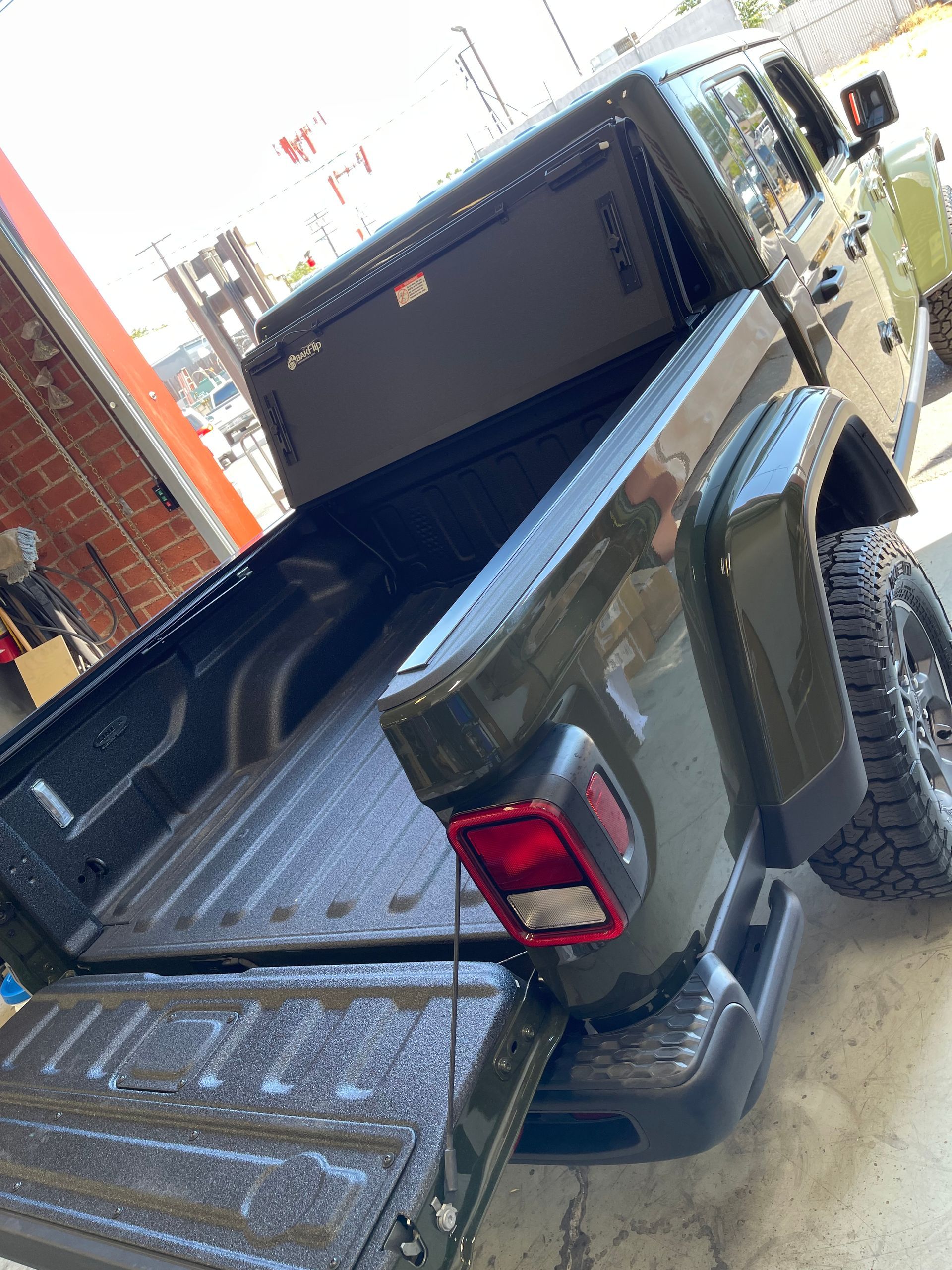 An olive green Jeep Gladiator pickup truck, viewed from the rear corner with the tailgate lowered.