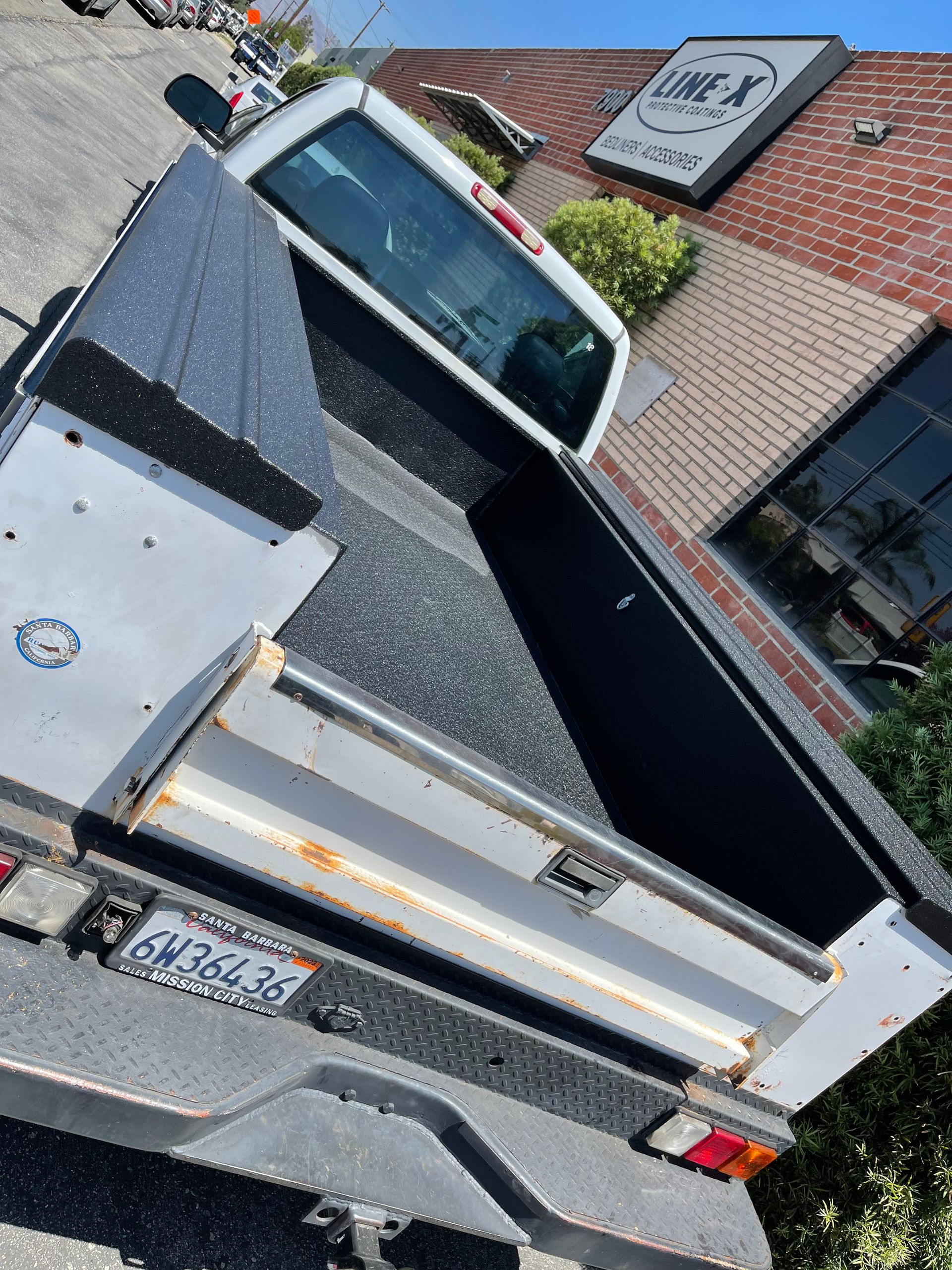 A white pickup truck bed with a fresh, black spray-on bedliner, parked outside a Line-X shop.