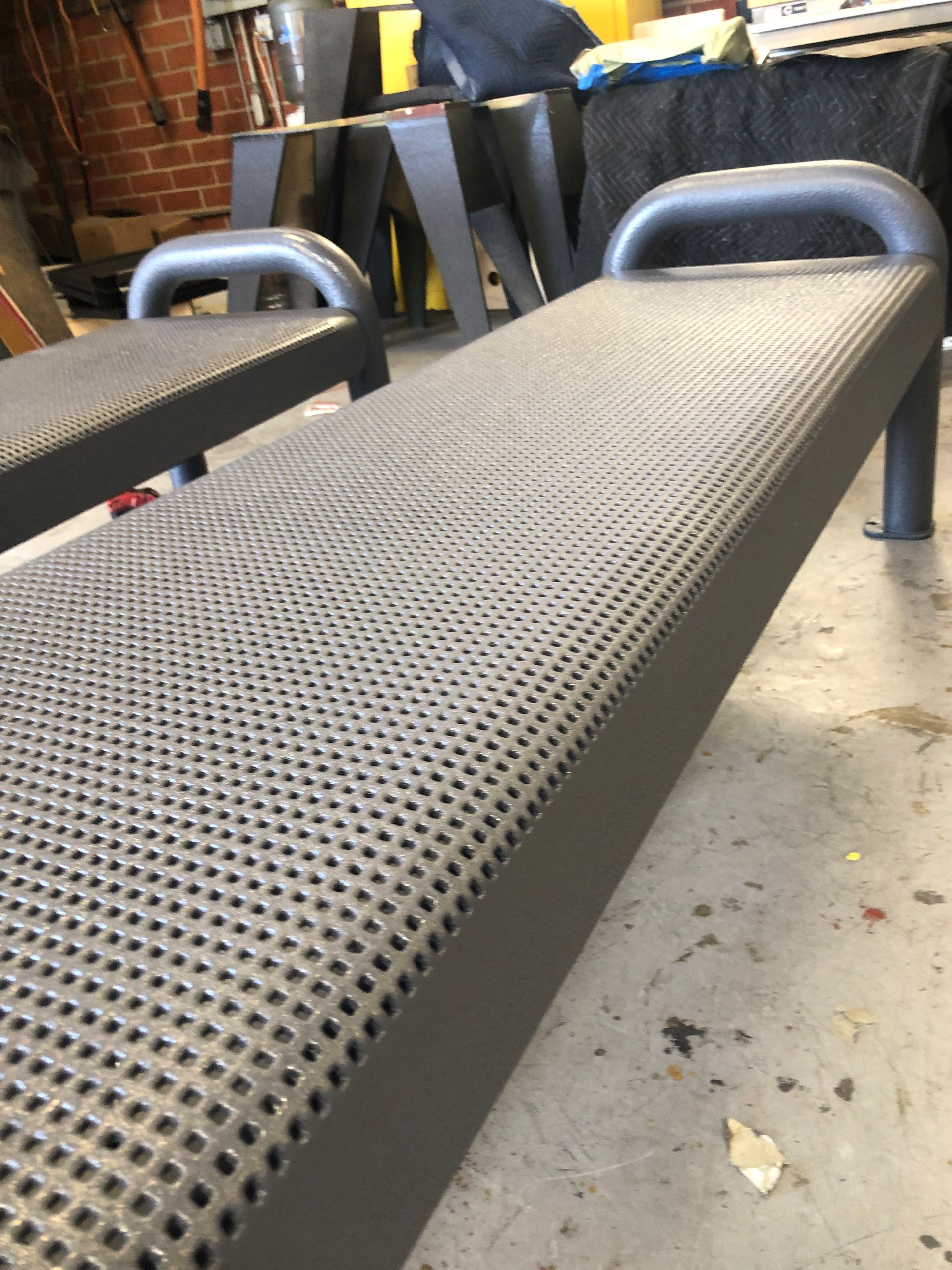 Two dark gray, perforated metal benches with curved end handles, positioned on a concrete floor in a workshop setting.