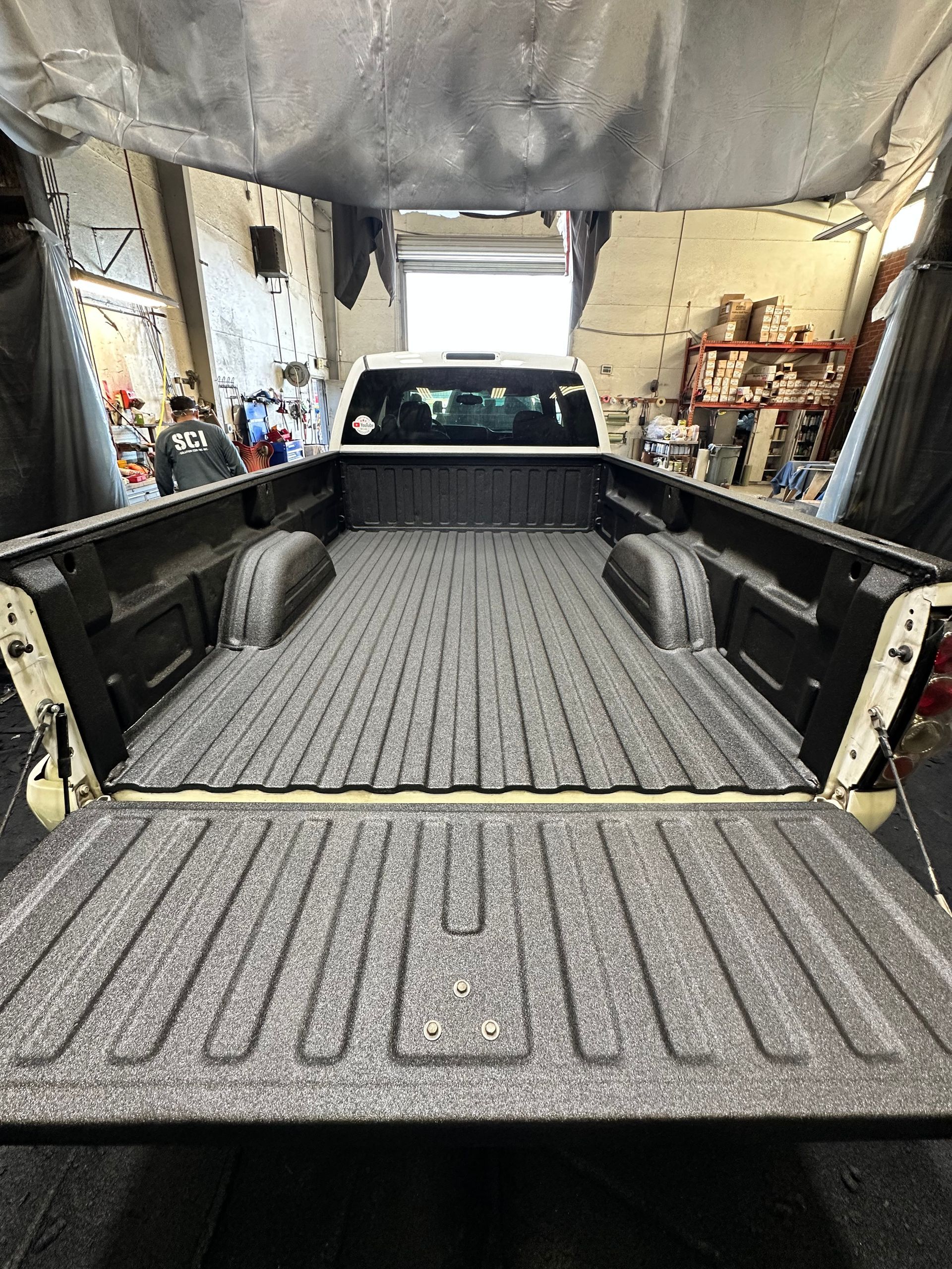 A truck bed featuring a newly applied black, textured spray-in bed liner inside an auto repair shop.