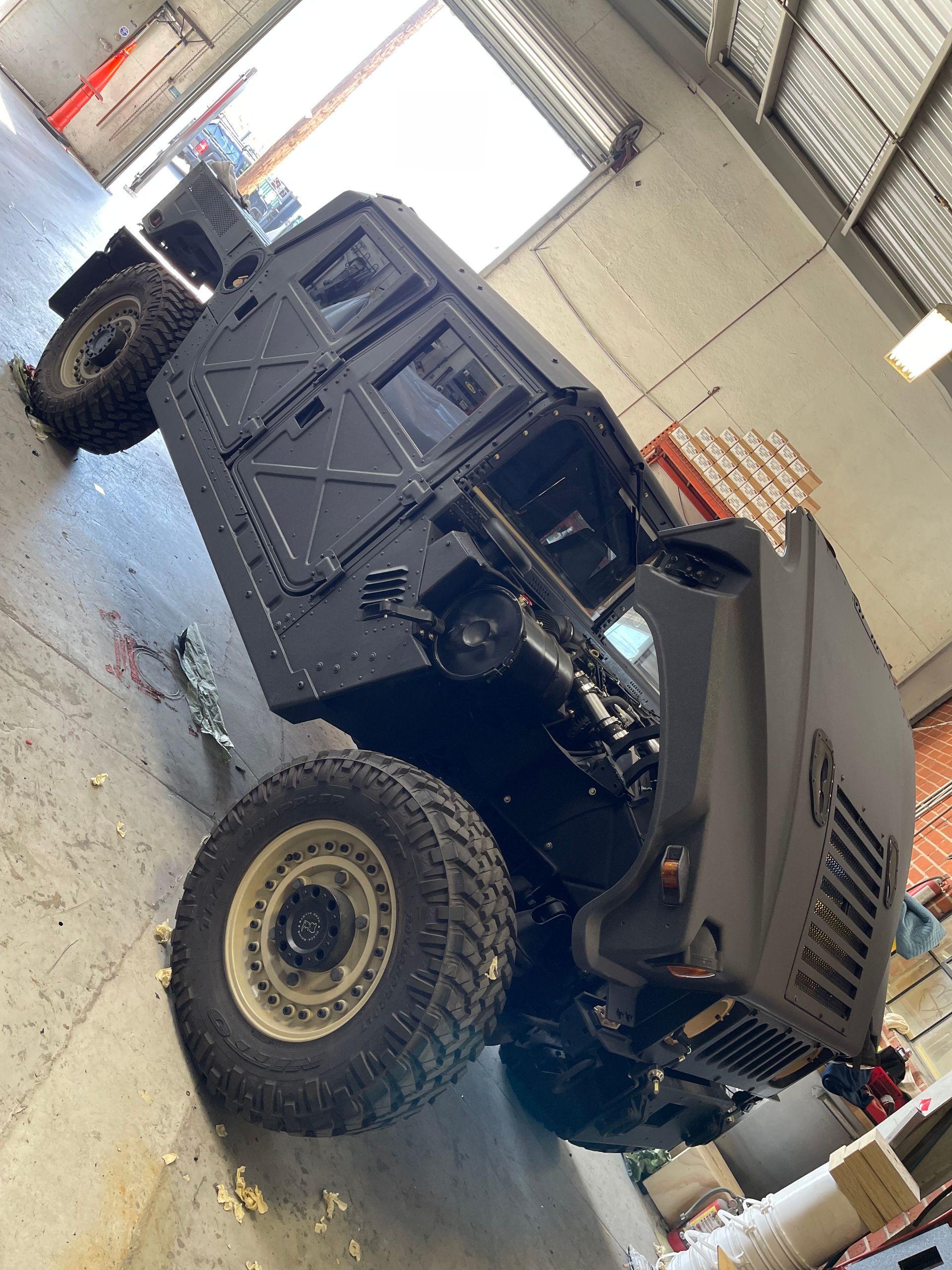 A matte black Humvee sits at an angle inside a workshop with a concrete floor and warehouse lighting.