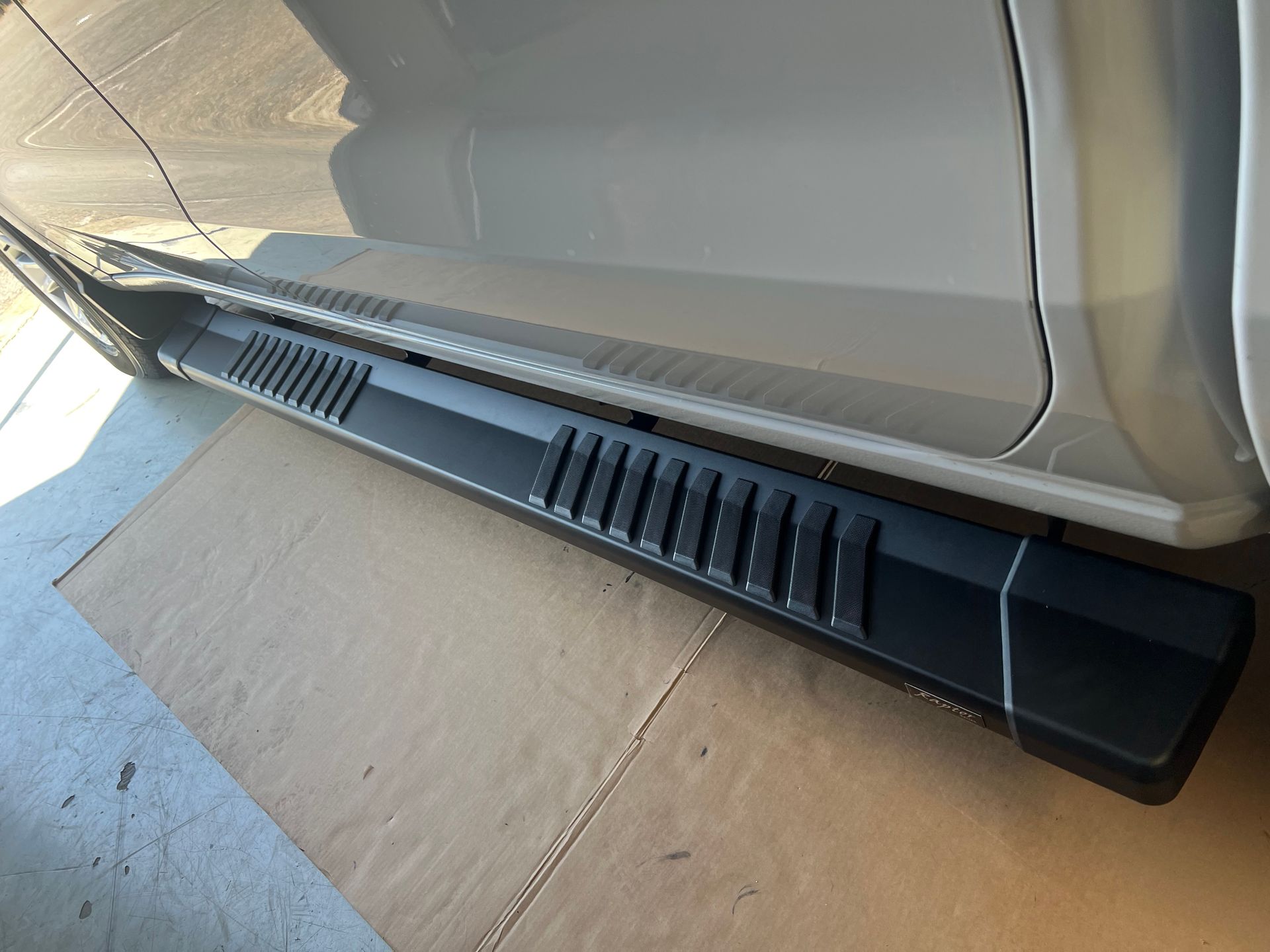 A matte black running board installed on the side of a light-colored vehicle, shown parked on a concrete surface.