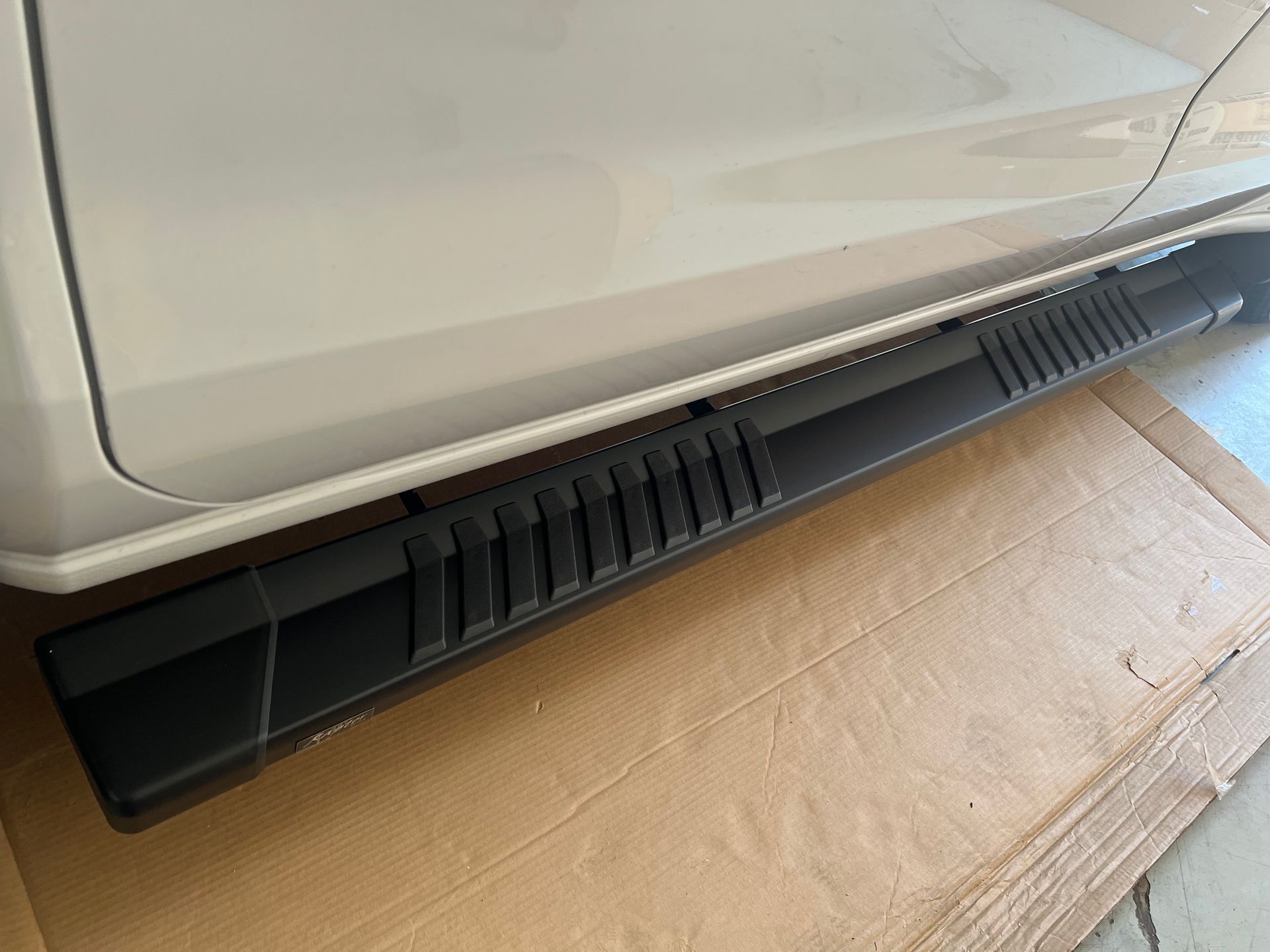 Black aftermarket running board installed on the side of a white vehicle, positioned over a cardboard surface.