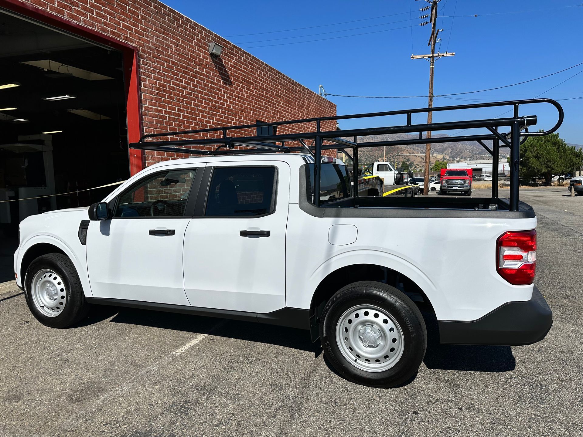 A white Ford Maverick pickup truck with a black utility rack, parked on a gravel lot next to a brick building.