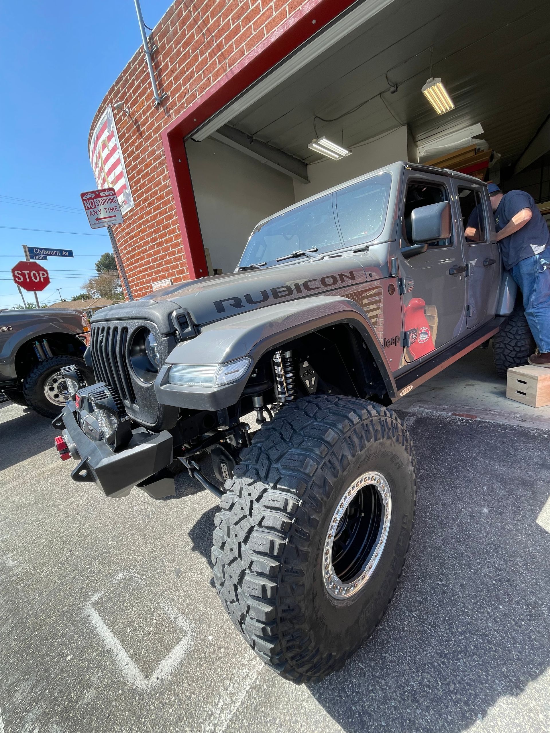 A gray Jeep Rubicon lifted on large off-road tires parked at a garage entrance under a clear blue sky.