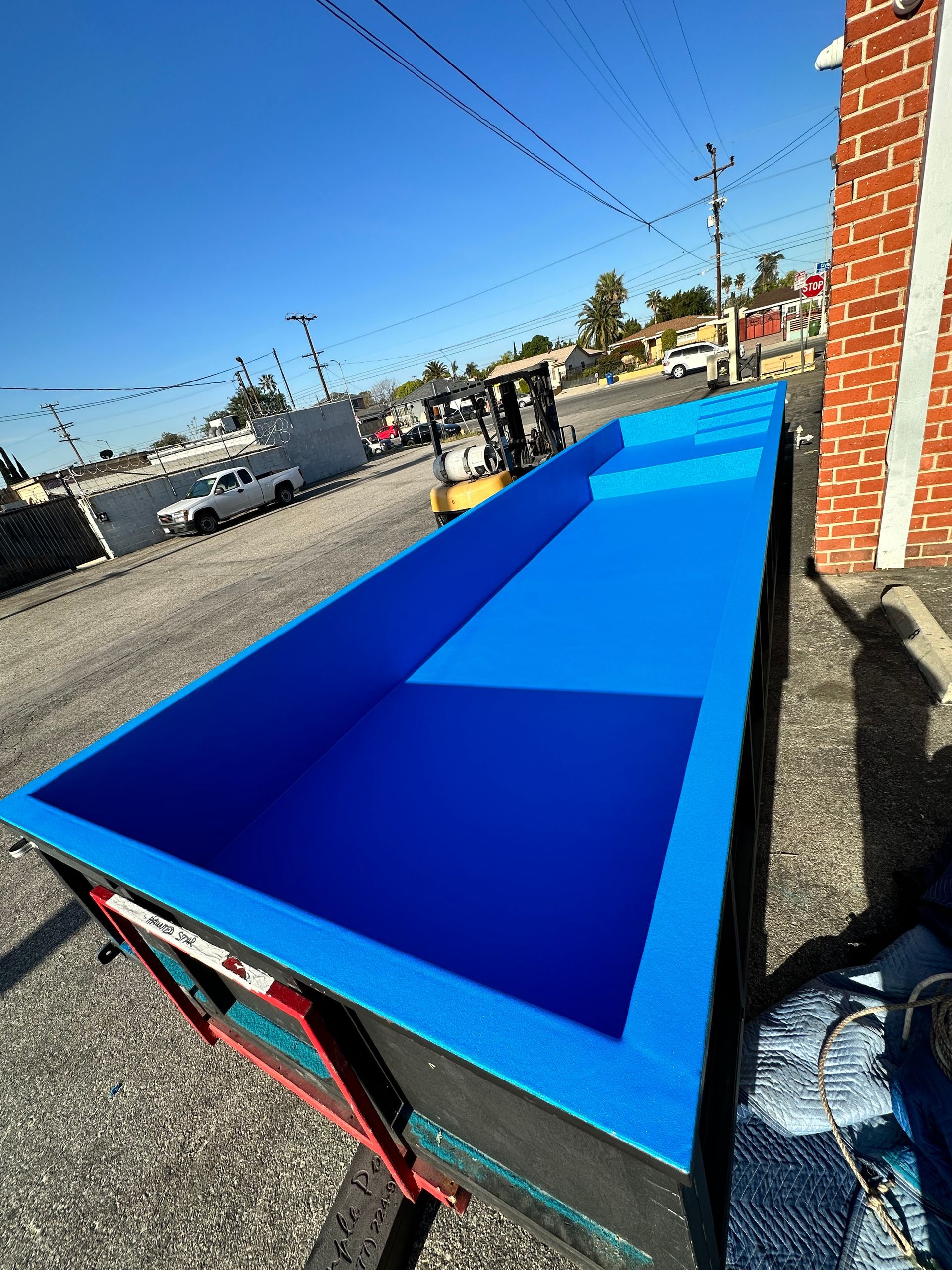 A long, bright blue fiberglass swimming pool shell sitting on a flatbed trailer in an outdoor storage yard.