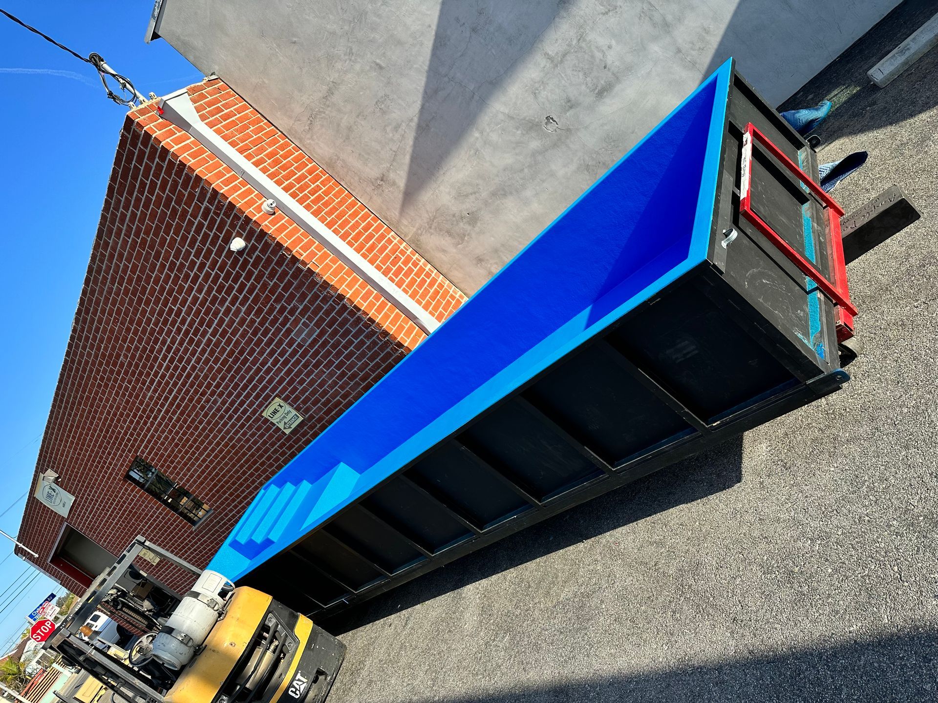 A yellow forklift lifts a long, empty, black-and-blue roll-off dumpster near a brick building.