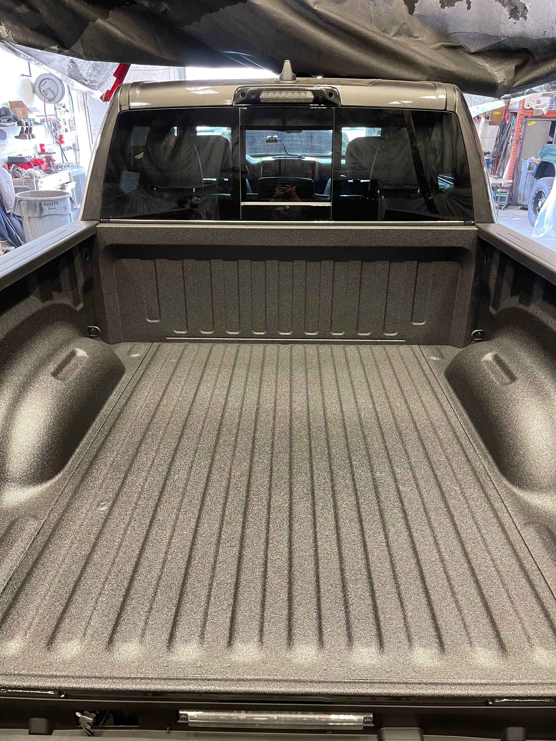 A truck bed with a grey textured spray-in bed liner, viewed from the rear, showing the truck's cabin window in the back.