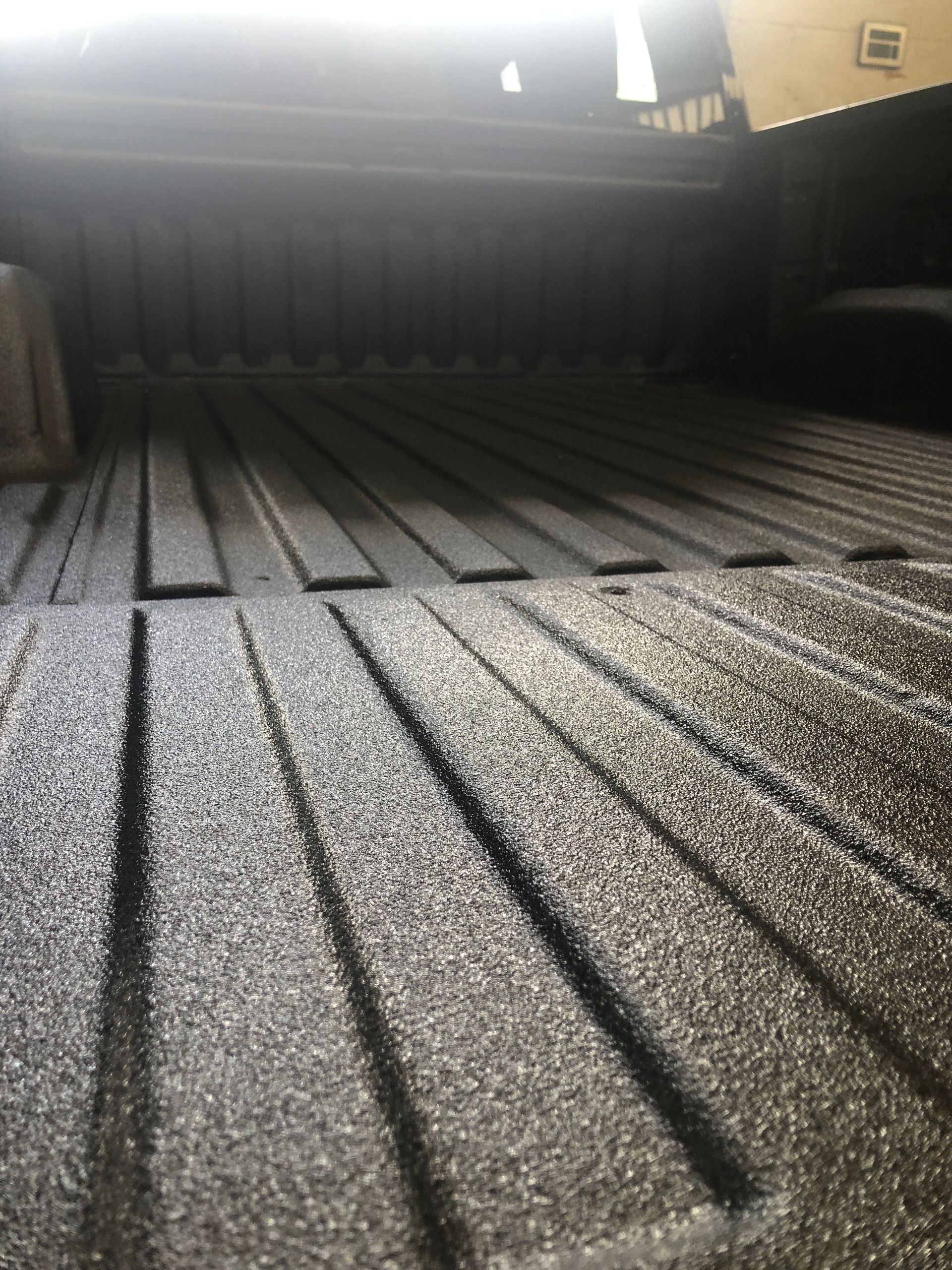 A close-up of a truck bed with a textured, black protective spray-in liner and raised longitudinal ridges.