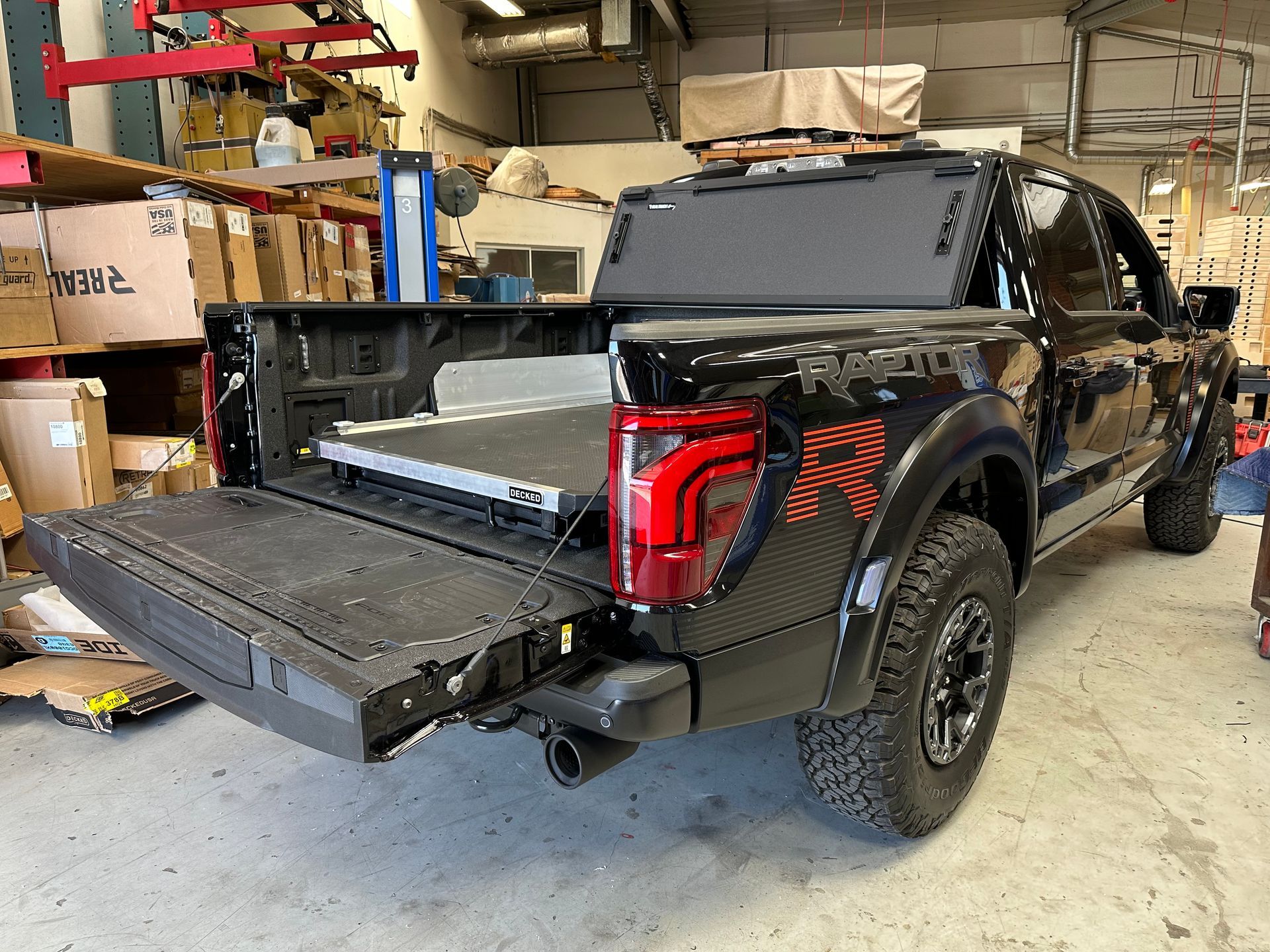 A black Ford Raptor truck parked in a garage with its tailgate down, revealing a storage drawer in the cargo bed.