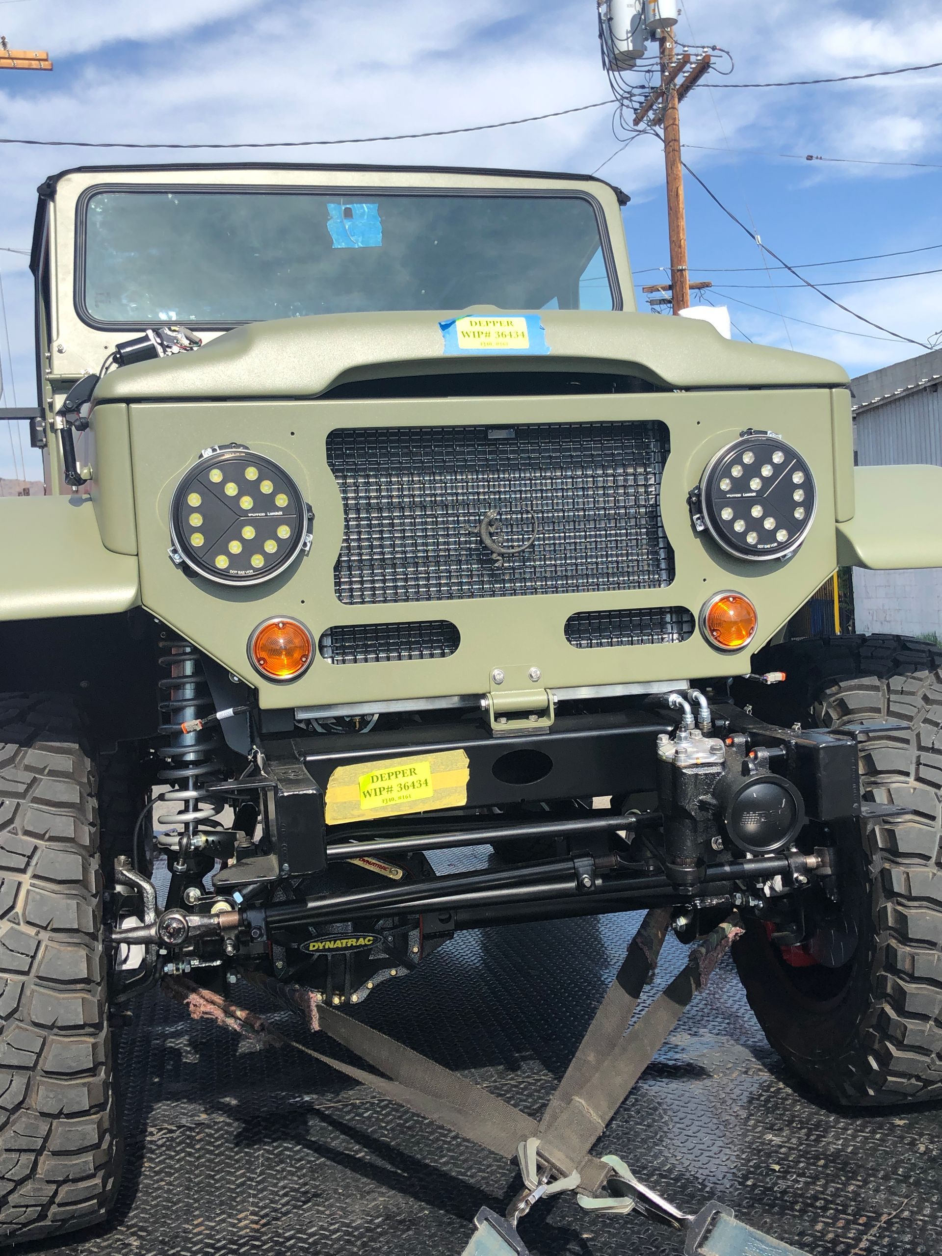 A matte olive drab off-road vehicle with large rugged tires and circular LED headlights, hitched and parked outdoors.