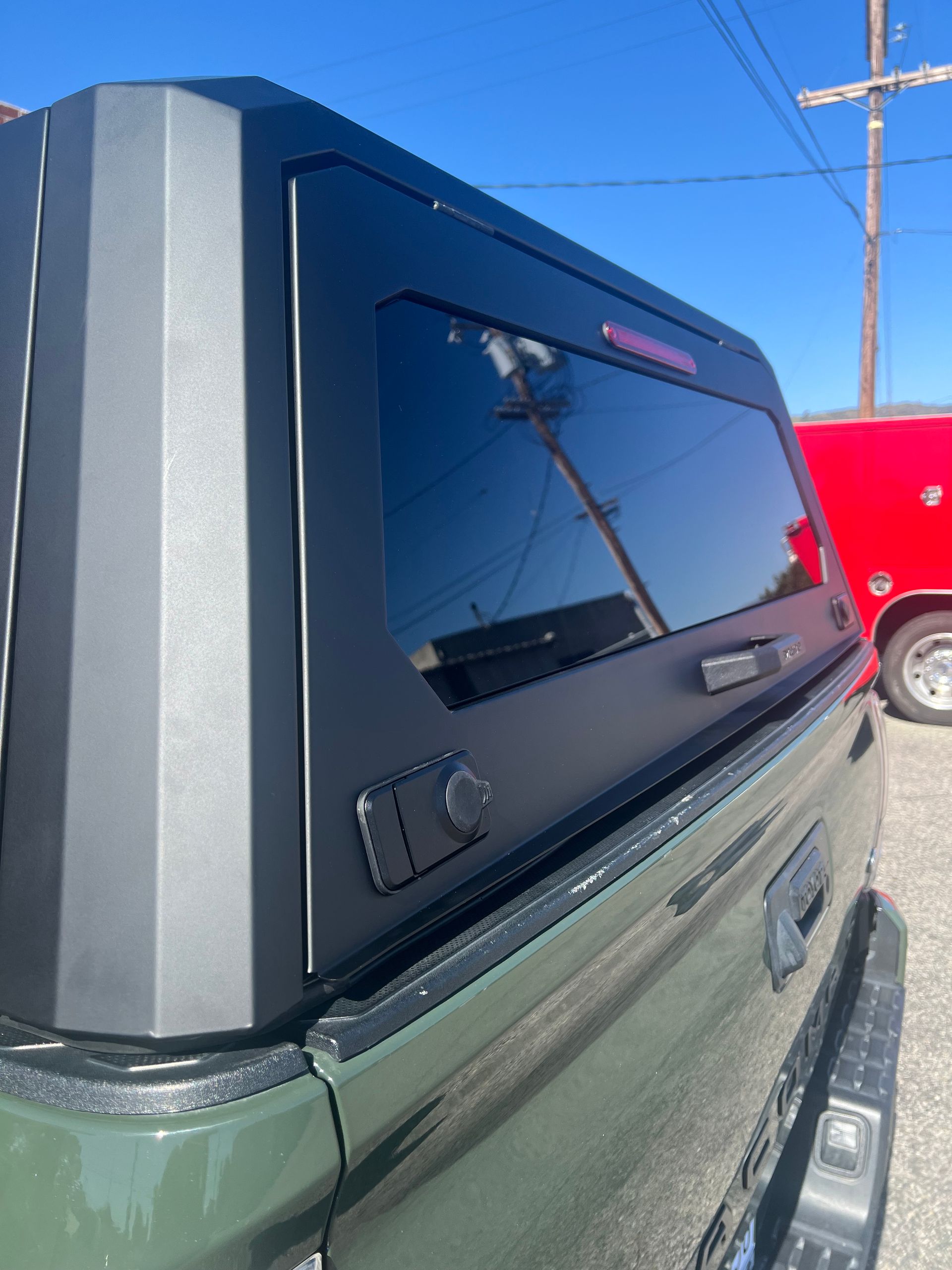 Close-up of a matte black truck camper shell installed on an army green pickup truck against a blue sky.