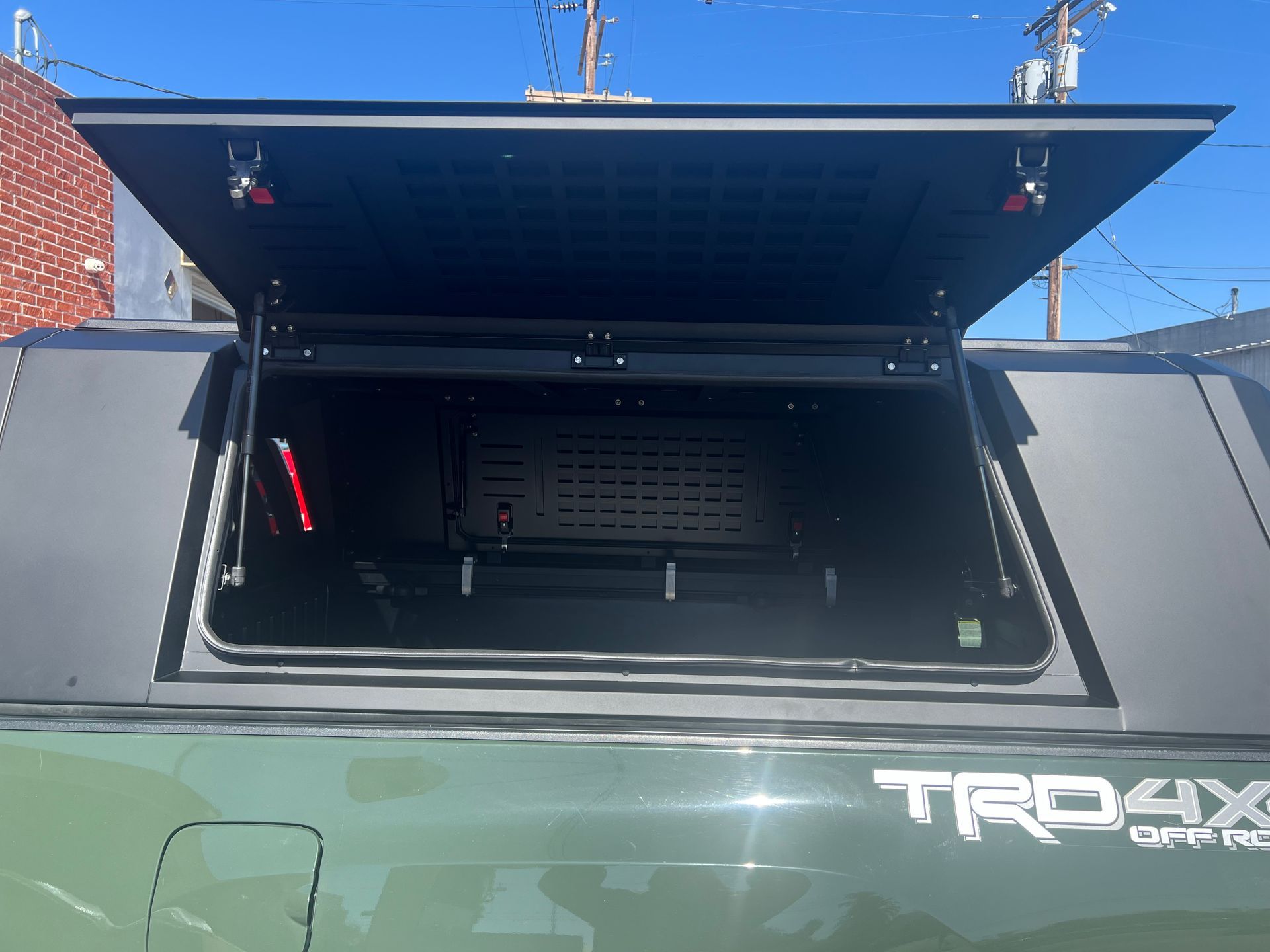 An open side hatch of a black truck bed topper mounted on a green TRD Off-Road truck against a clear blue sky.