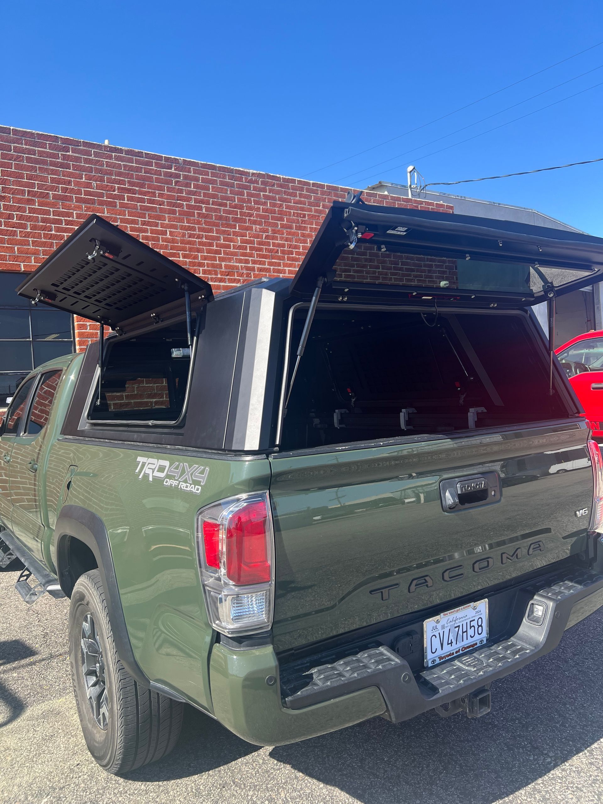 Army green Toyota Tacoma parked outside, featuring a black aftermarket truck topper with side and rear hatches opened.