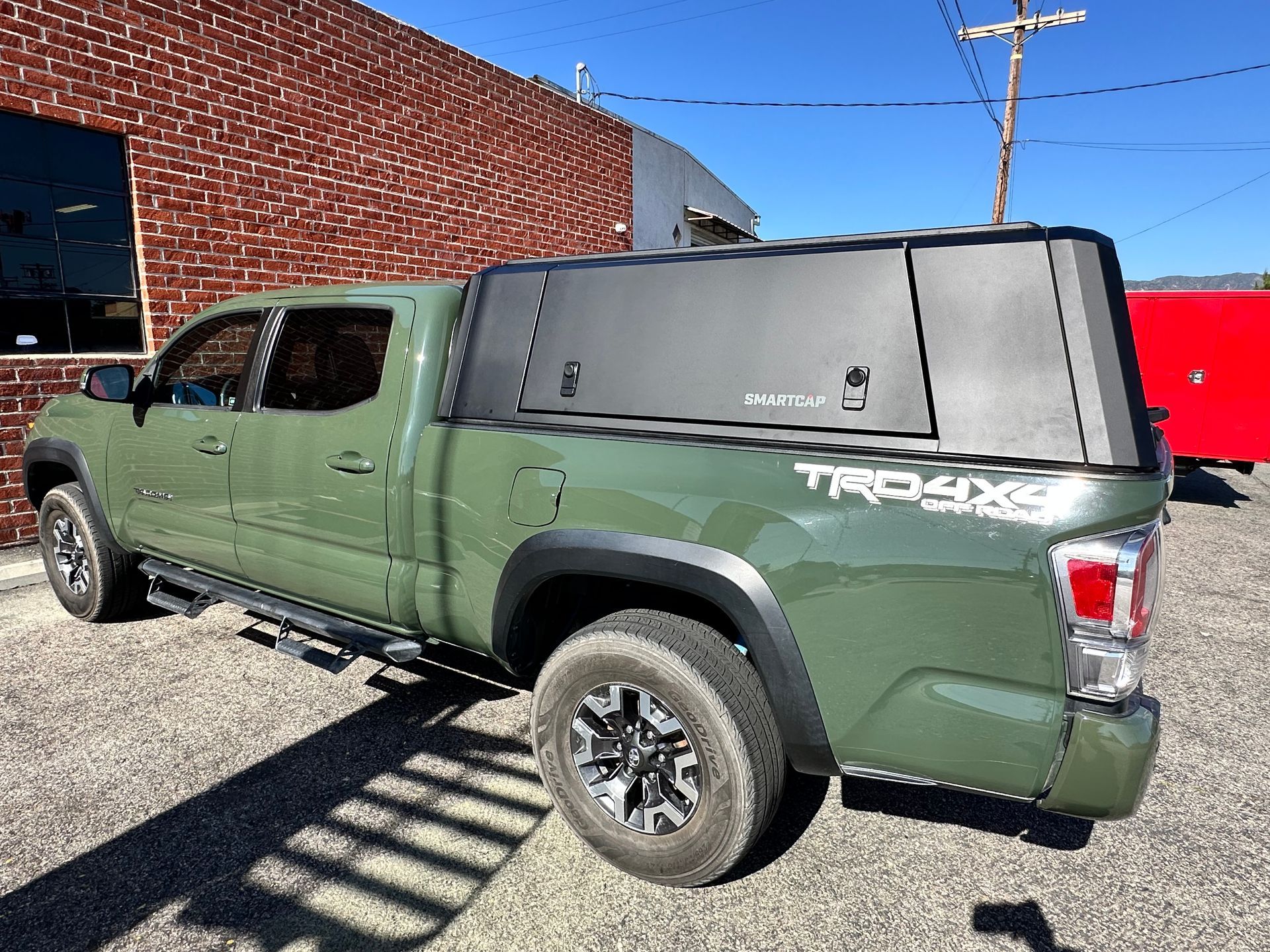 Army green Toyota Tacoma pickup truck with a black camper shell parked on a gravel lot in front of a brick building.