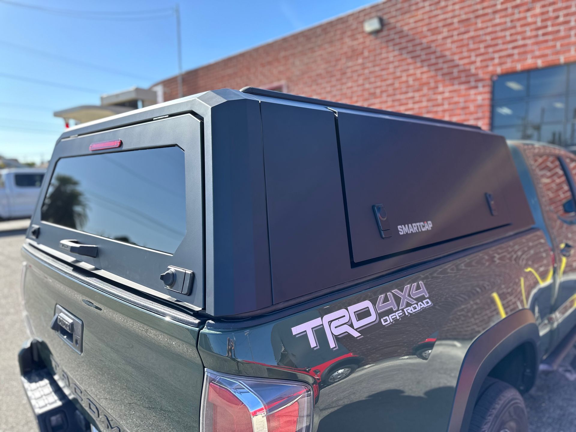A black, angular truck topper installed on a green TRD 4x4 Toyota Tacoma parked outside a brick building.