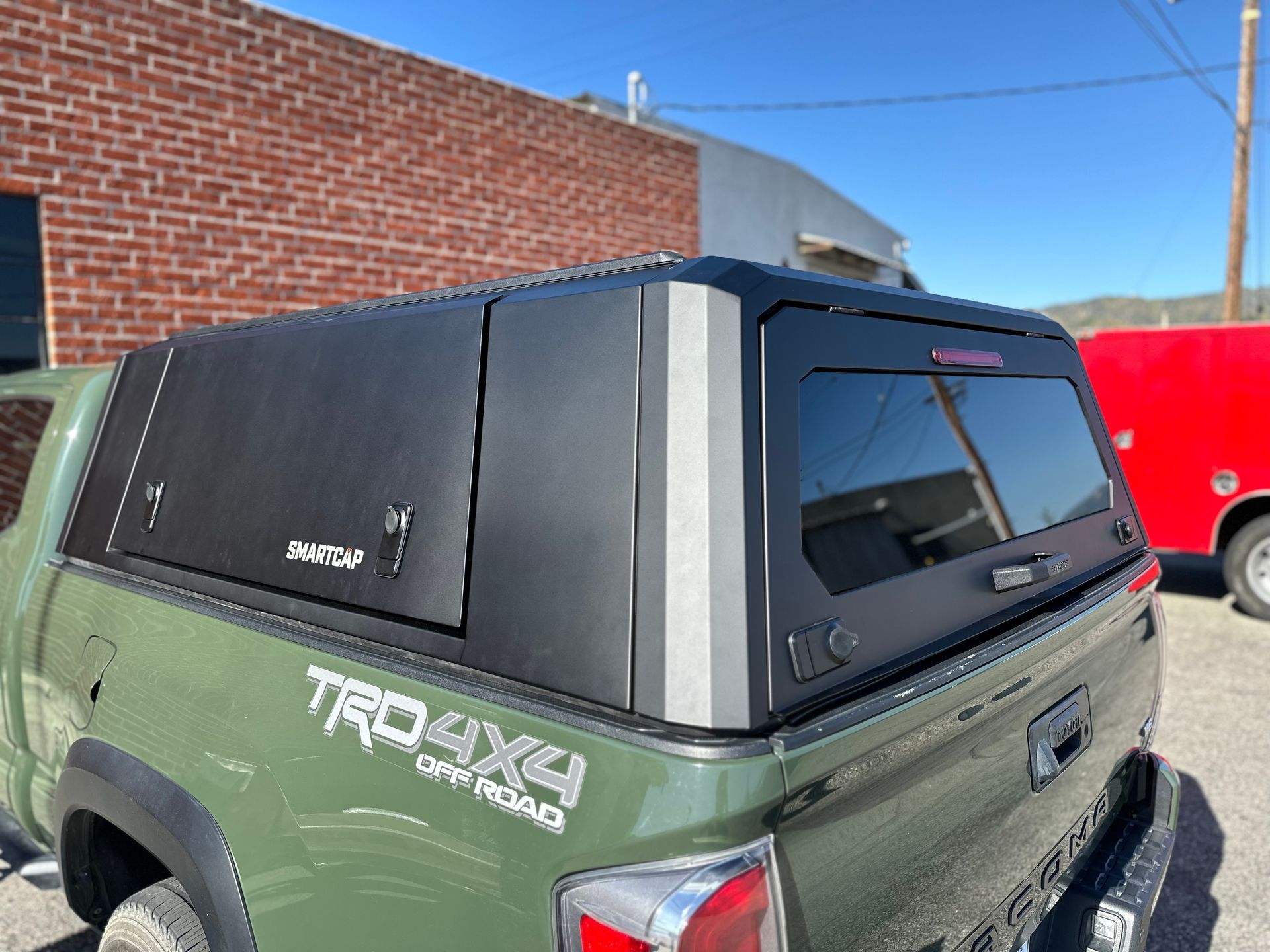 An olive green Toyota Tacoma truck equipped with a black metal camper shell, parked outside a brick building.