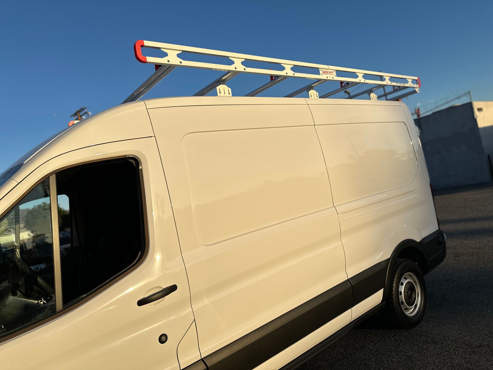 A white commercial cargo van parked outdoors with a silver metal ladder rack mounted on the roof.