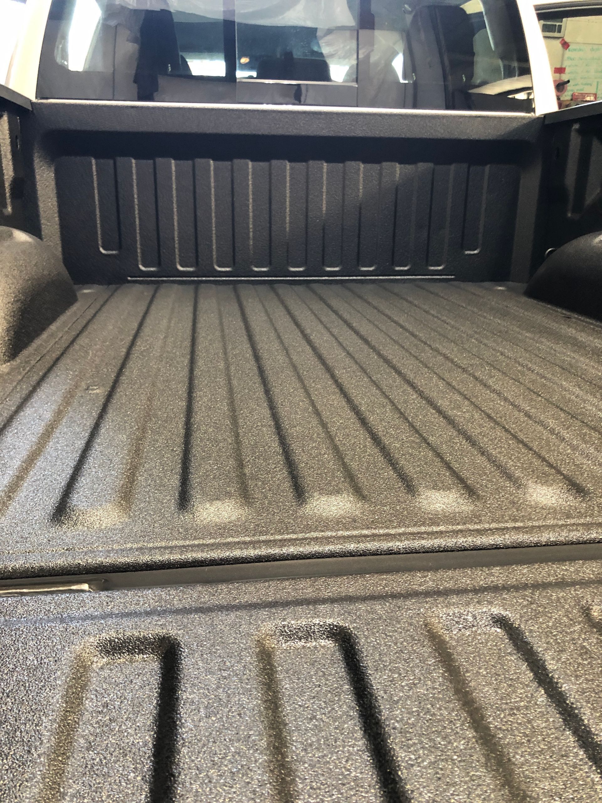 A view into the truck bed of a vehicle, featuring a dark, textured spray-in bed liner covering the floor and walls.