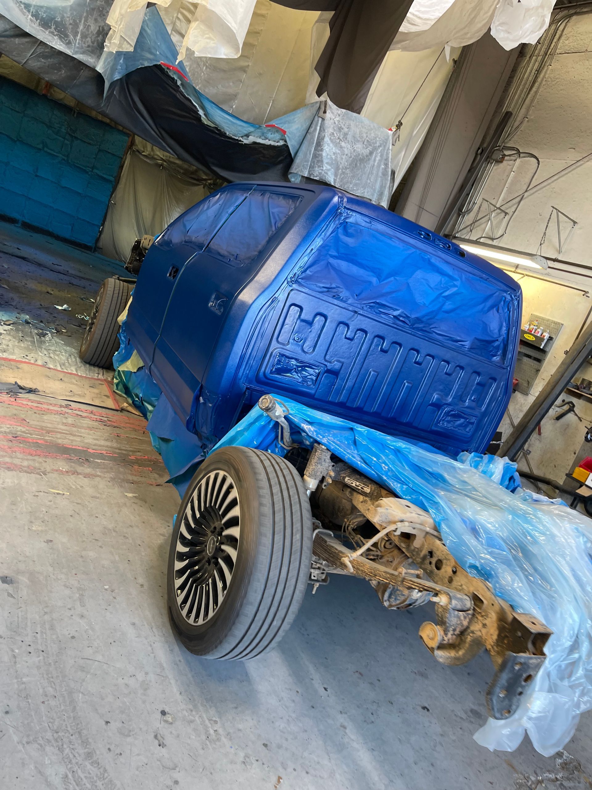 A blue vehicle cab, partially wrapped in blue plastic, sits in a garage with one visible wheel and exposed chassis.