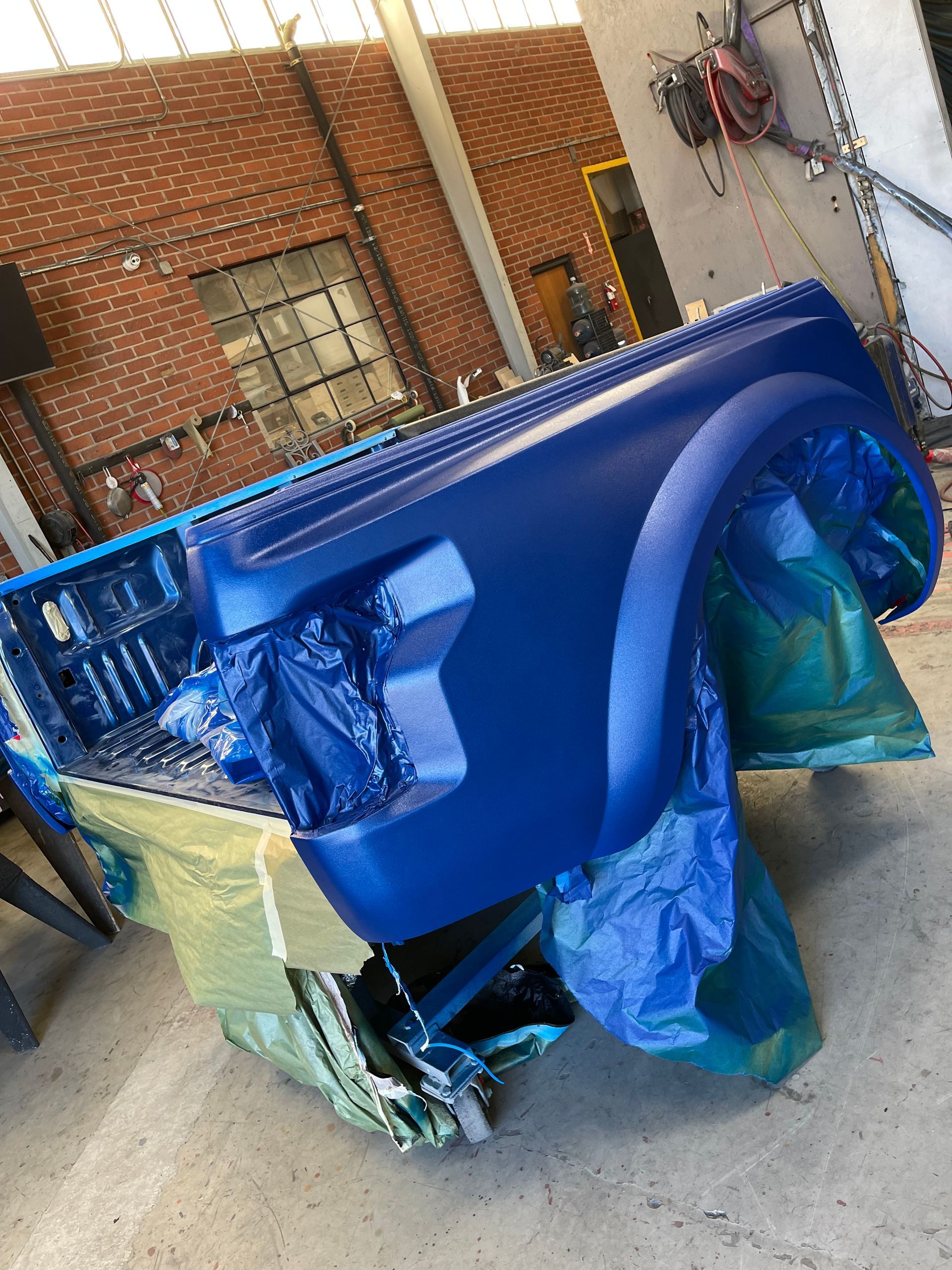 A freshly painted, bright blue truck bed sits on a stand in a workshop, partially covered by masking plastic.