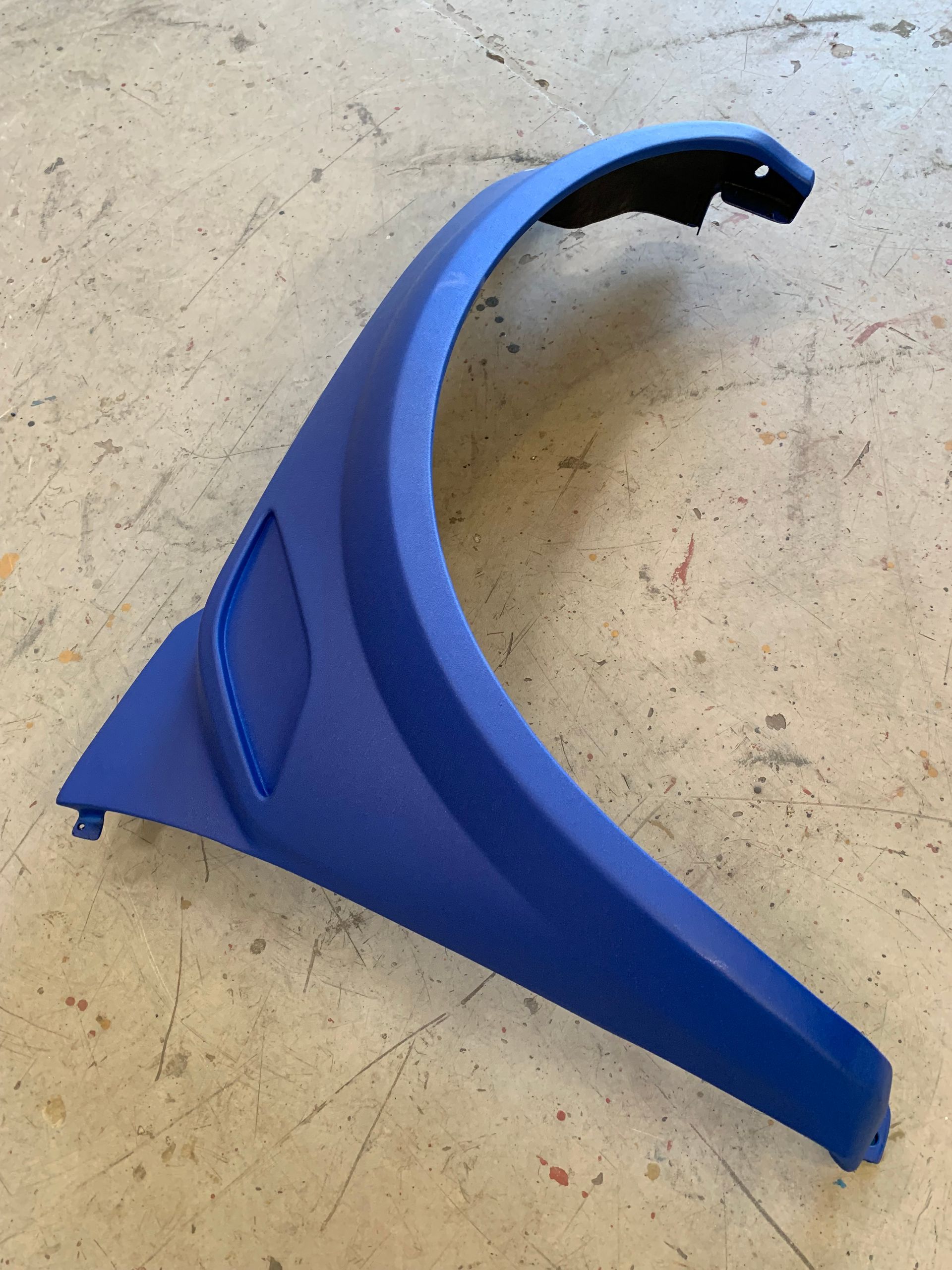 A blue, curved vehicle fender flare or trim piece resting on a concrete floor.