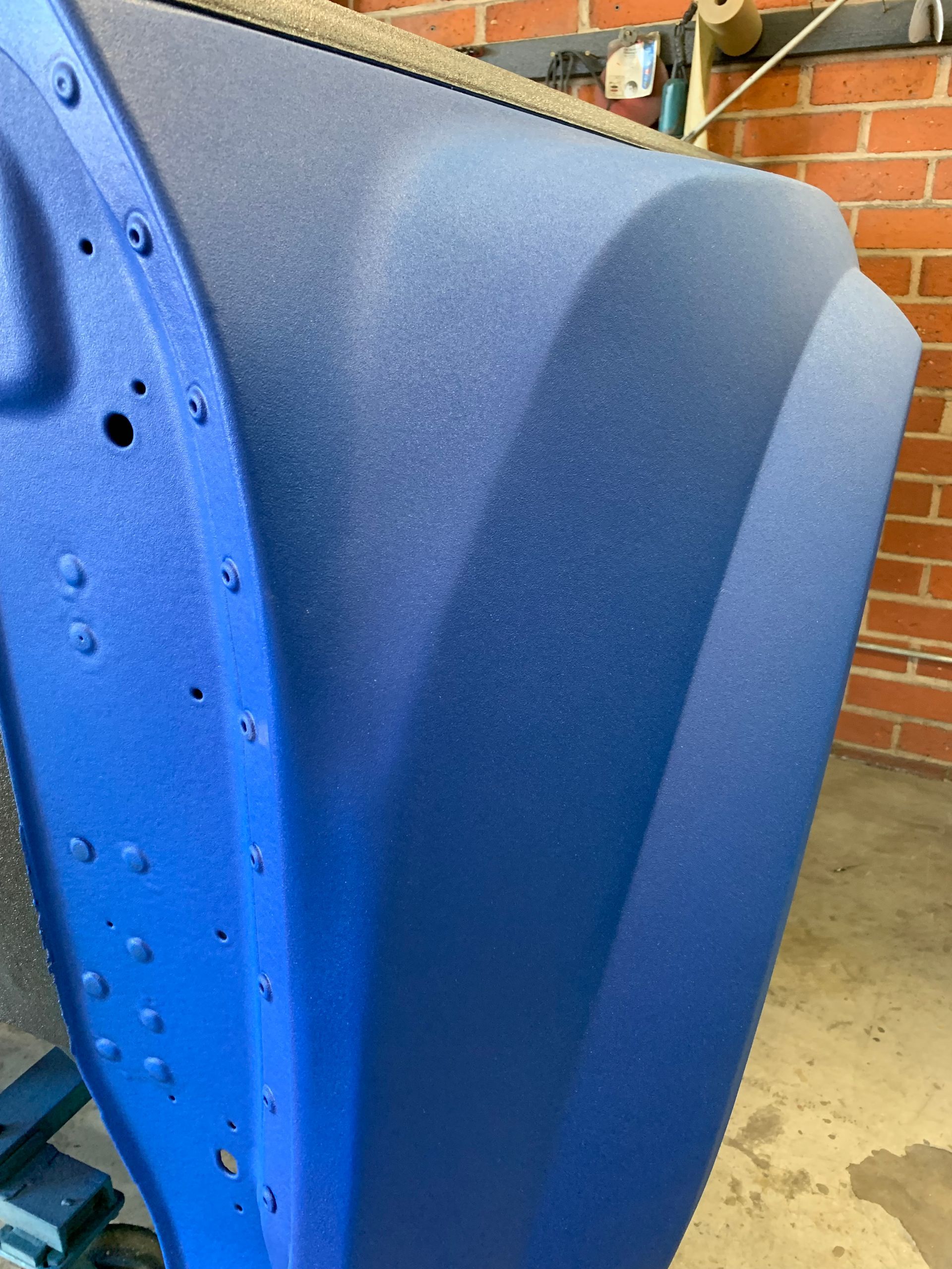 A car fender coated in a bright blue, textured, grit-finish paint, shown in a workshop against a brick wall.