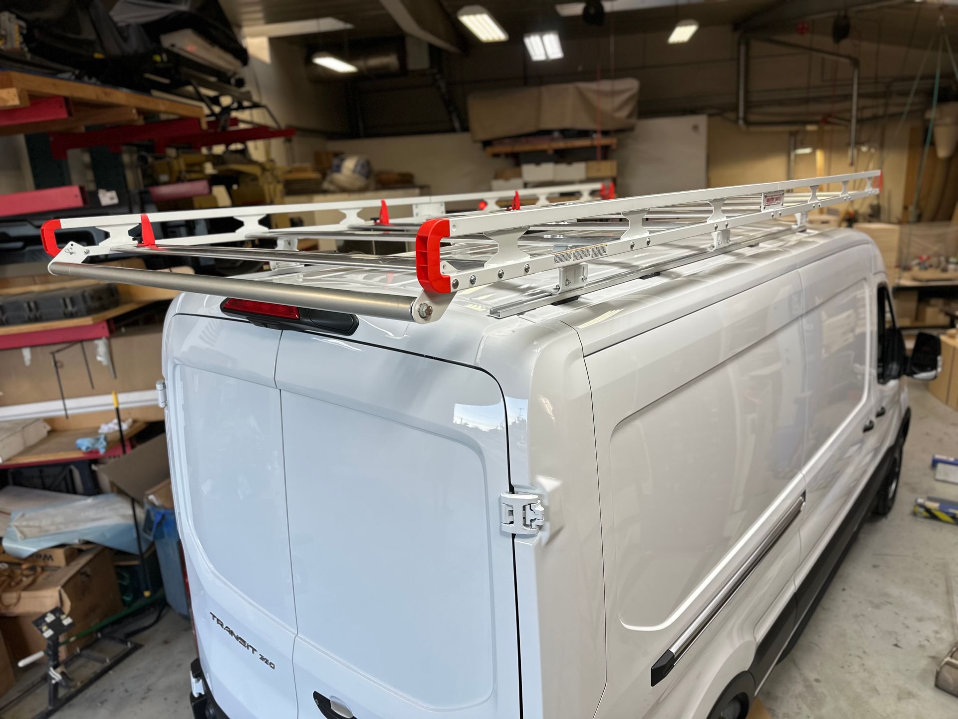 A white cargo van parked indoors, equipped with a large aluminum roof rack featuring bright red safety brackets.