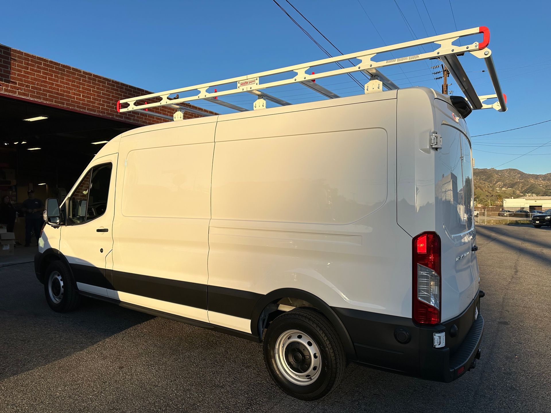 A white Ford Transit commercial cargo van with a metal roof ladder rack parked on an asphalt lot.