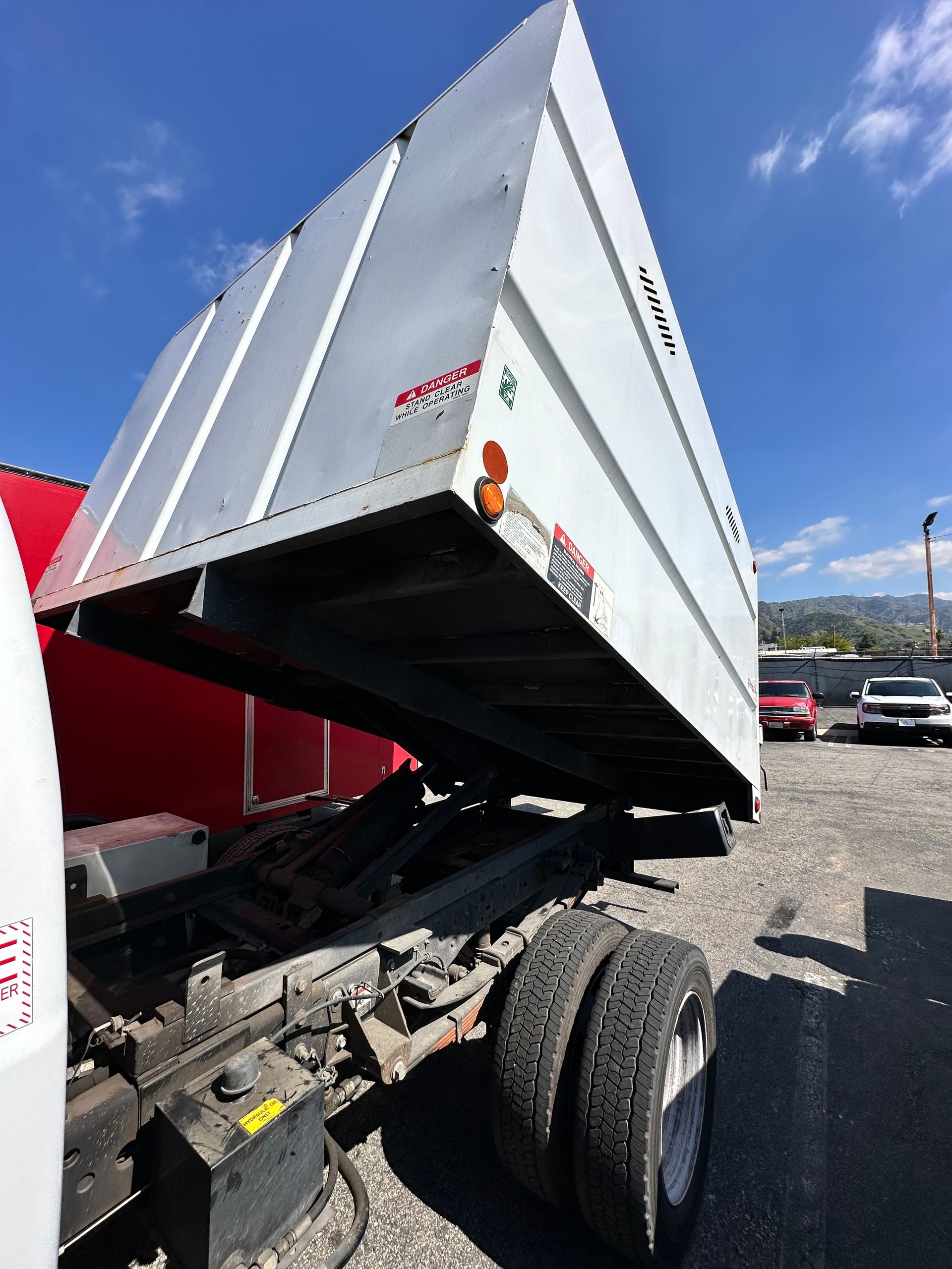 A white dump truck with its bed fully raised in an outdoor lot on a sunny day.