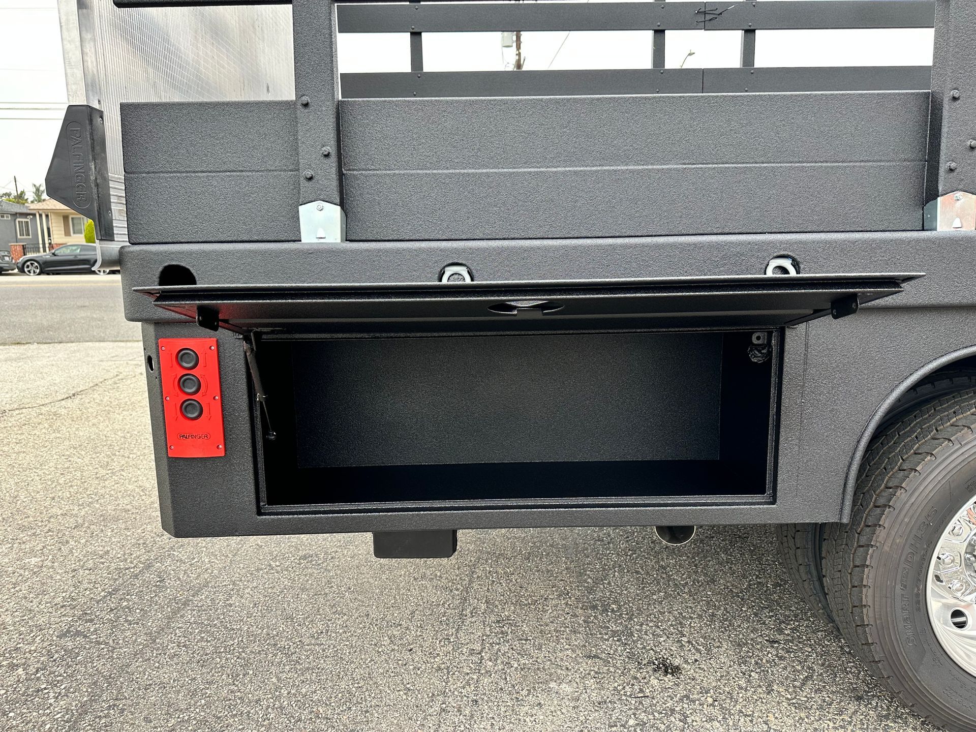 A side view of a black truck bed with an open storage compartment revealing a dark interior and red electrical outlets.
