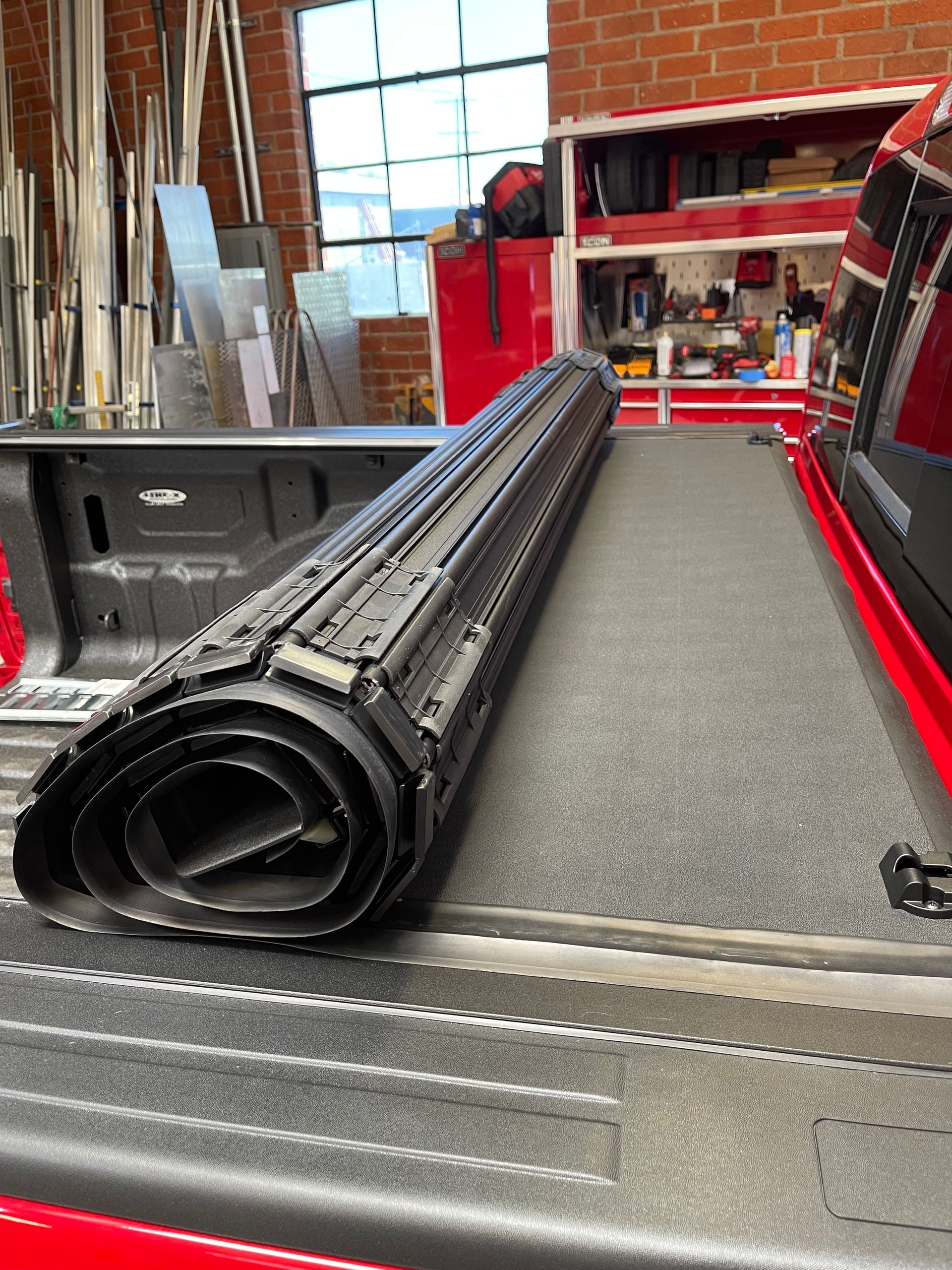 A black rolled-up truck bed cover sits on the surface of a red truck bed inside a garage workshop.