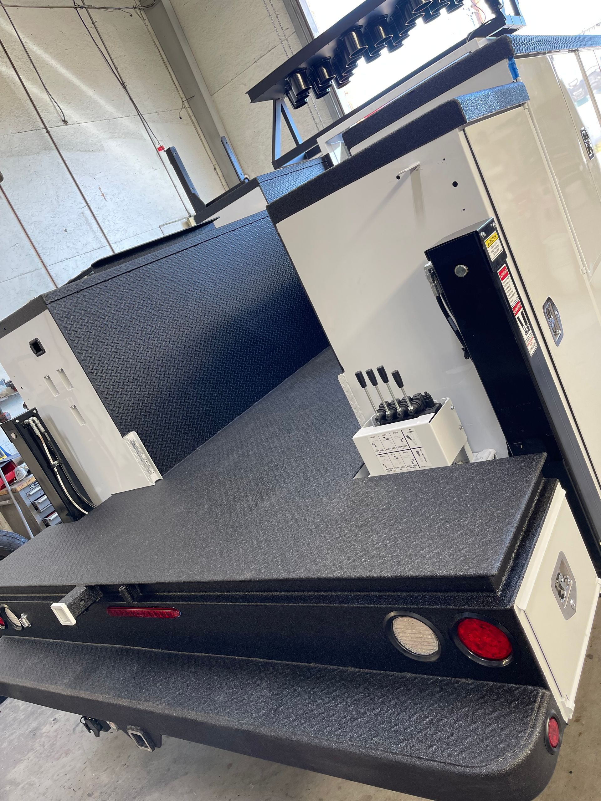 A flatbed truck body with a black diamond-plate headache rack, side storage compartments, and hydraulic control levers.