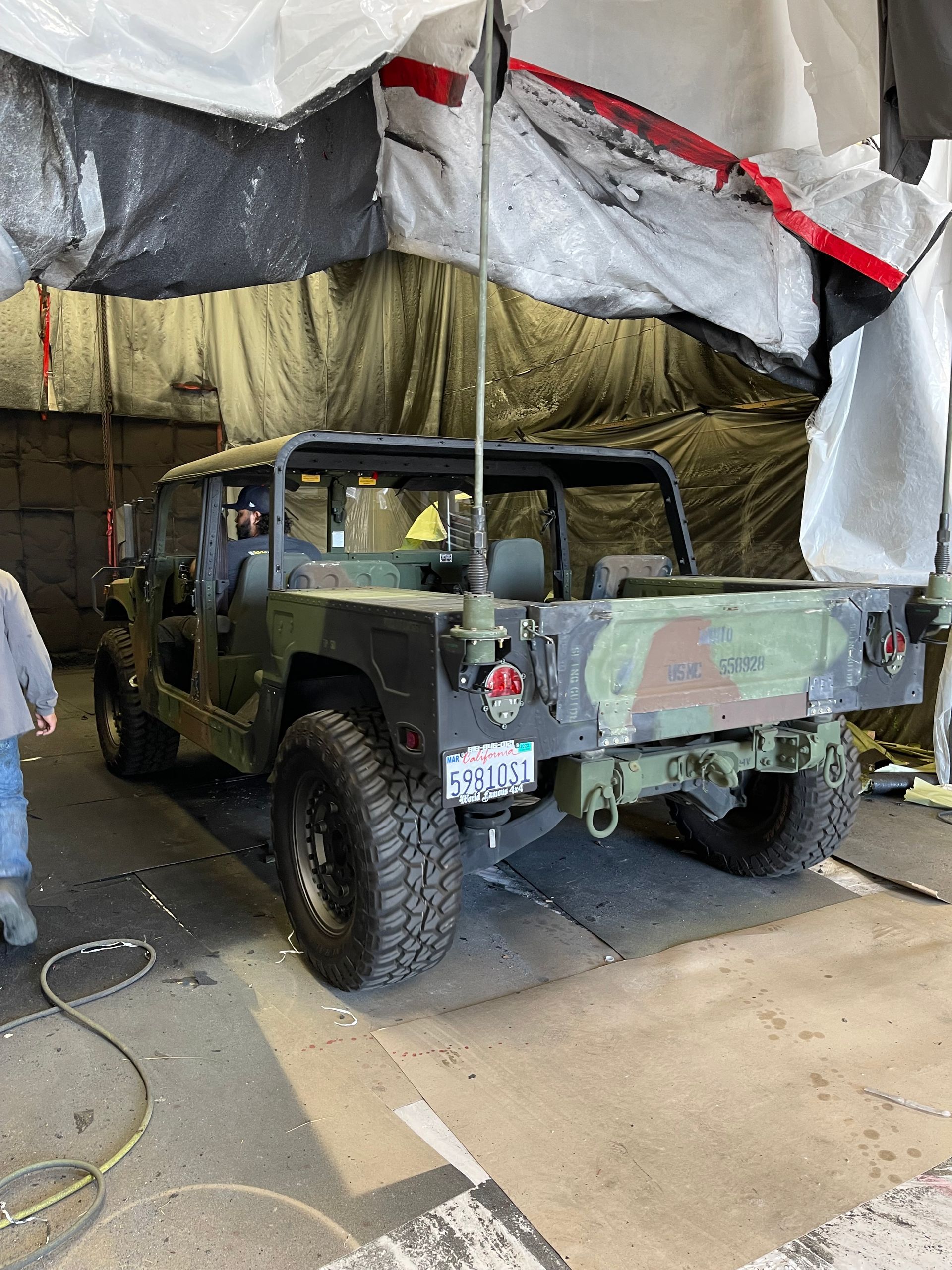 A camo-patterned Humvee parked inside a garage, partially covered by a white tarp, with a person standing to the left.