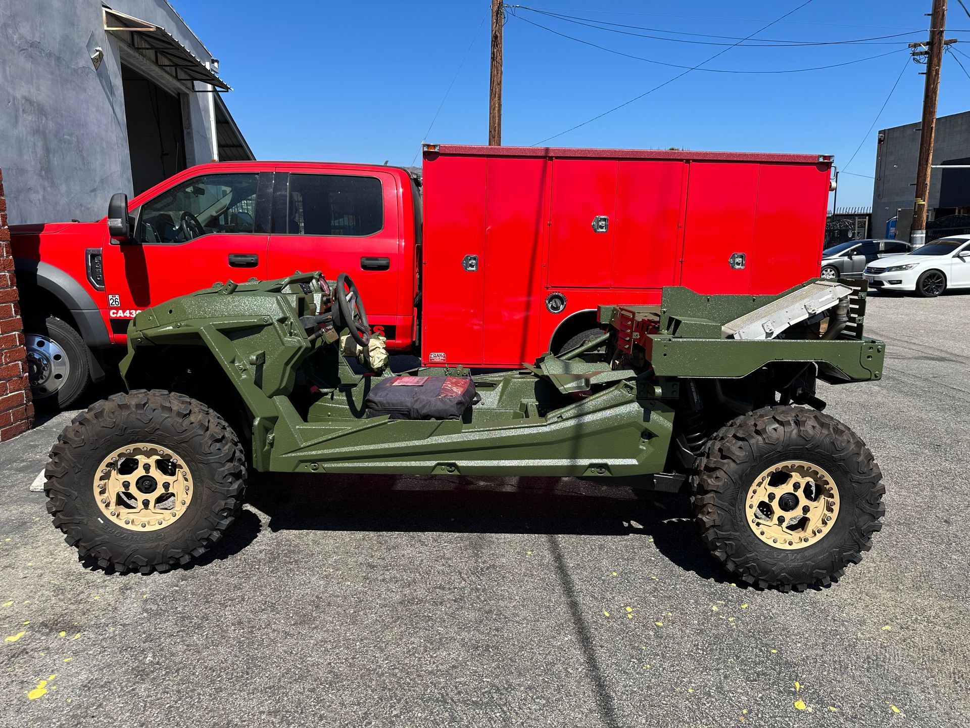 A green, open-frame military-style vehicle parked in front of a red utility truck on a paved outdoor lot.