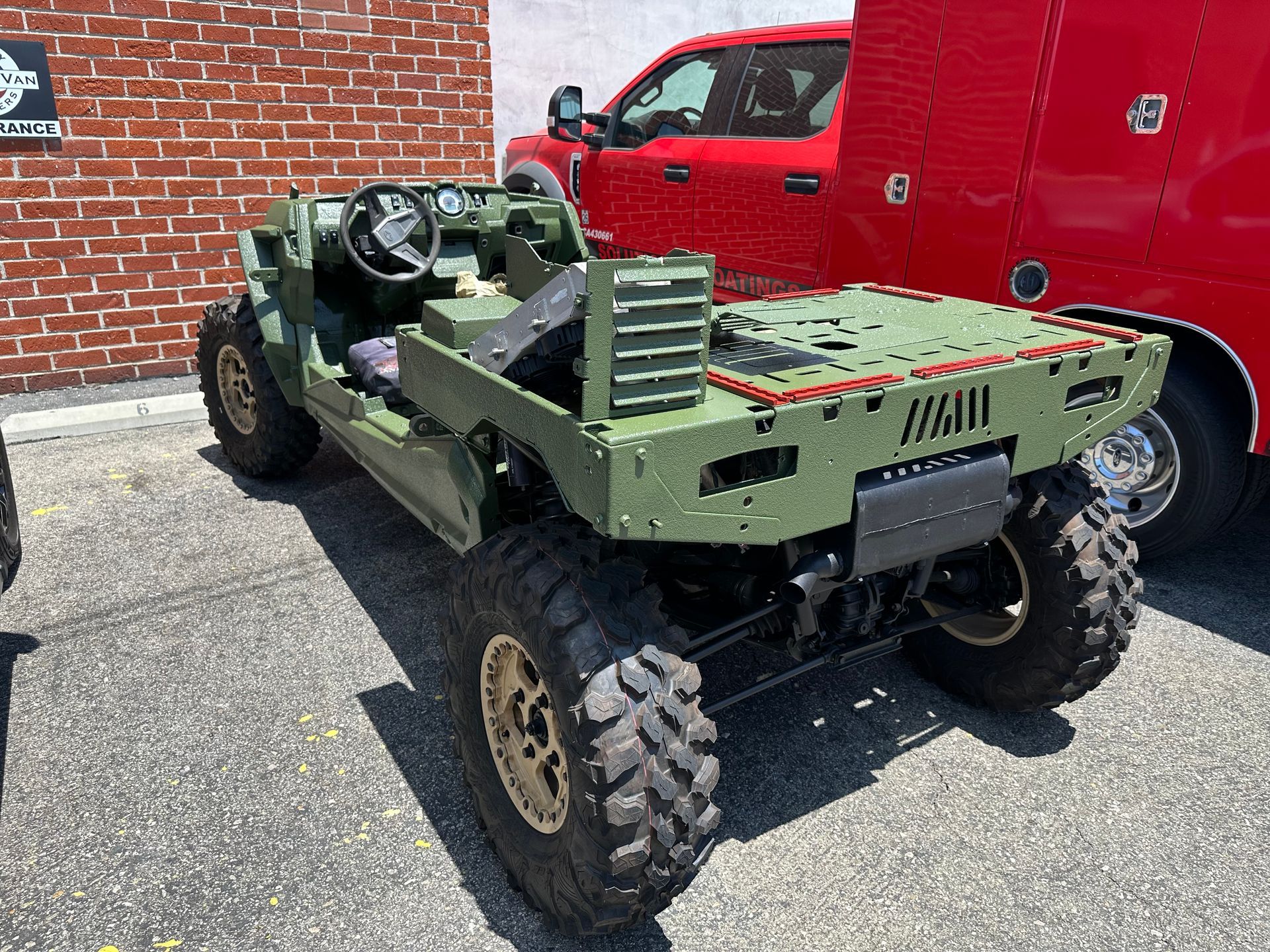 A green military-style tactical vehicle parked on asphalt in front of a red utility truck and a brick wall.