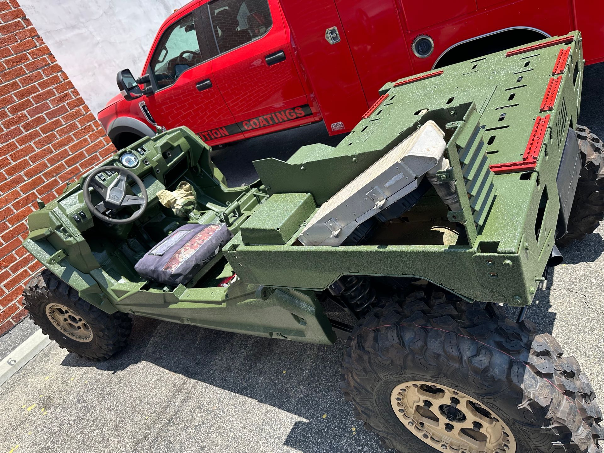 A green, tactical off-road military vehicle parked on asphalt next to a red truck and a brick wall.