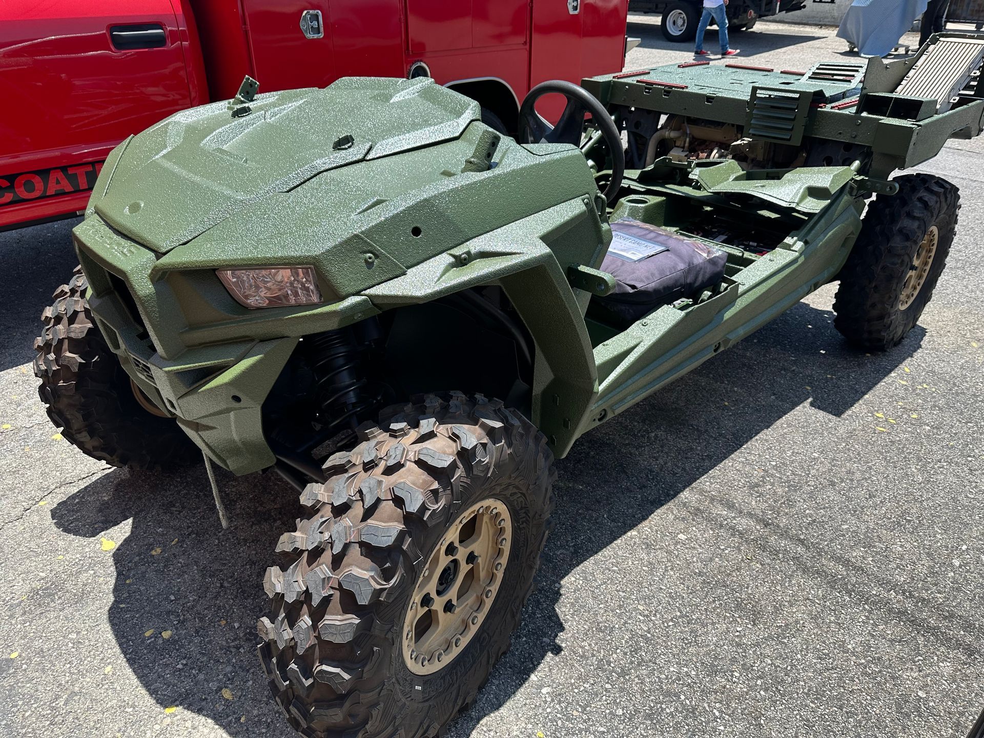 A green, military-style all-terrain vehicle parked on asphalt next to a red truck.