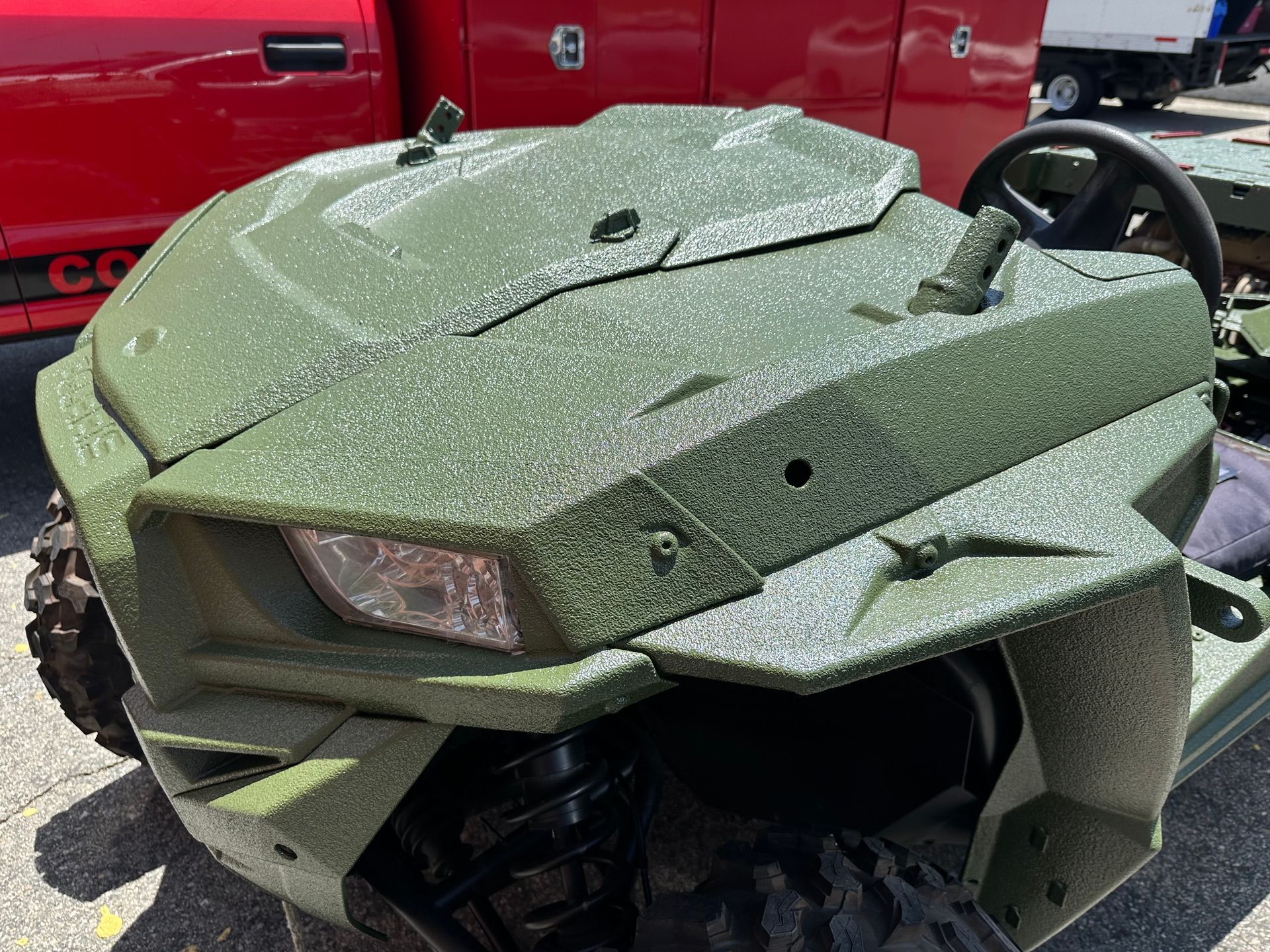A front view of an olive green, textured, military-style ATV hood with a headlight, parked near a red utility truck.