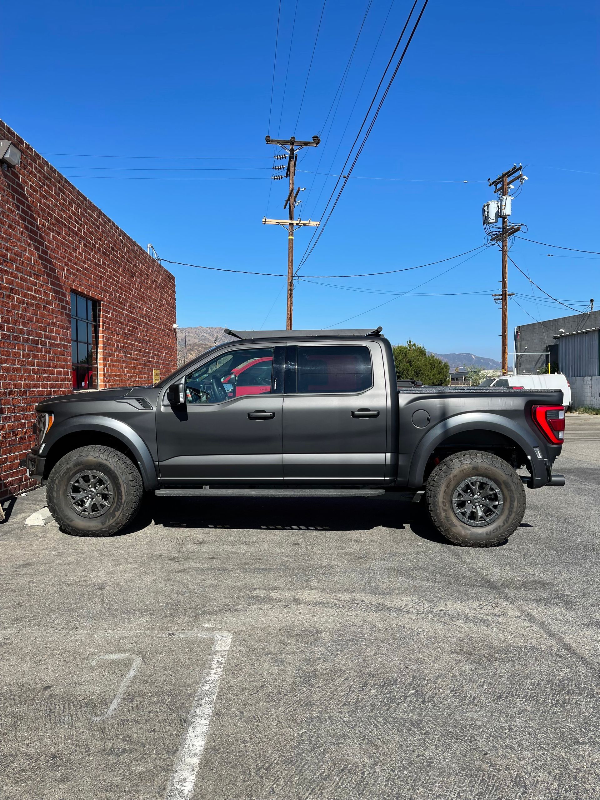 A dark gray Ford F-150 Raptor pickup truck parked on a gravel lot next to a brick building under a clear blue sky.
